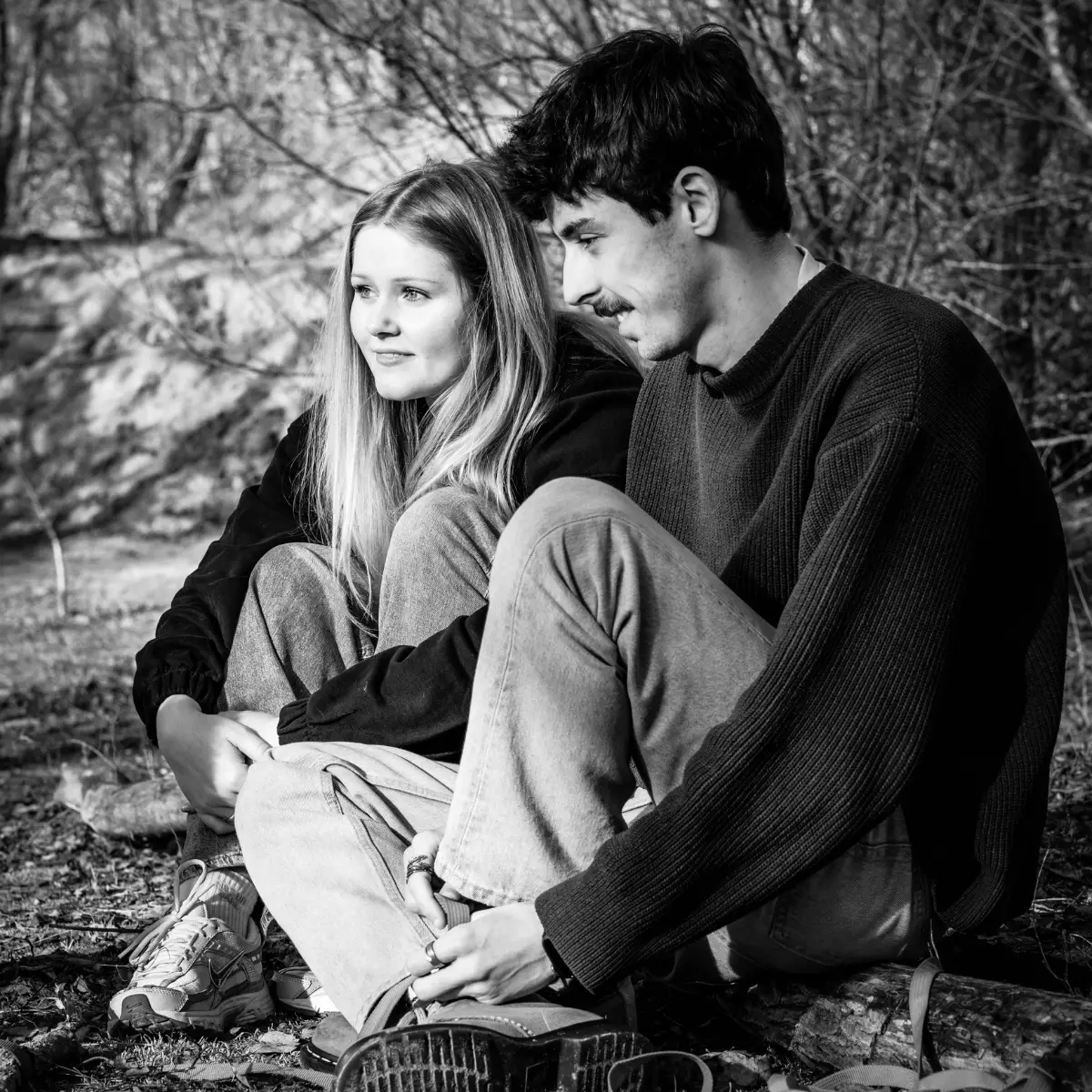 Zwart-witfoto van een jonge man en vrouw die buiten op de grond zitten, ontspannen en glimlachend, met de vrouw die haar knieën omarmt en de man die zijn schoenen vastpakt, omringd door bomen en bladeren.