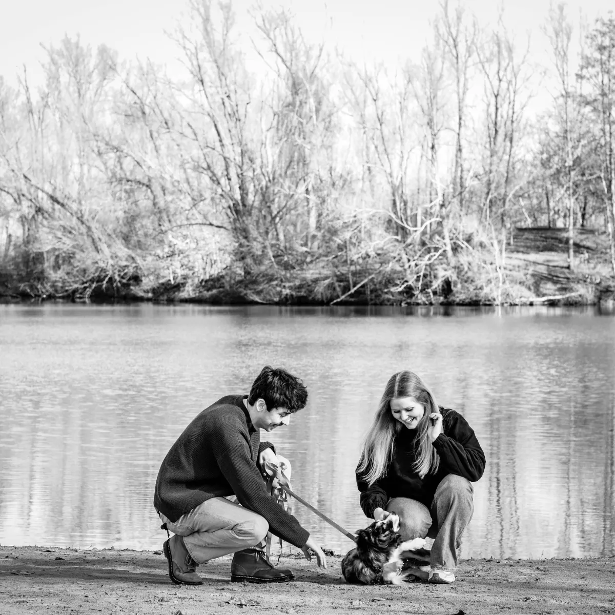 Jongeman en jonge vrouw knielen bij het water aan een meer en spelen met een kleine hond aan een riem op de oever, omringd door bomen zonder bladeren.
