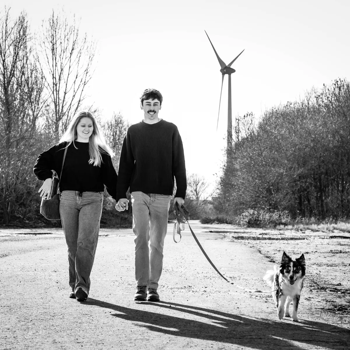 Een jong koppel loopt hand in hand over een onverharde weg met een blije hond aan een riem voor hen uit, omringd door bomen en een windmolen op de achtergrond.