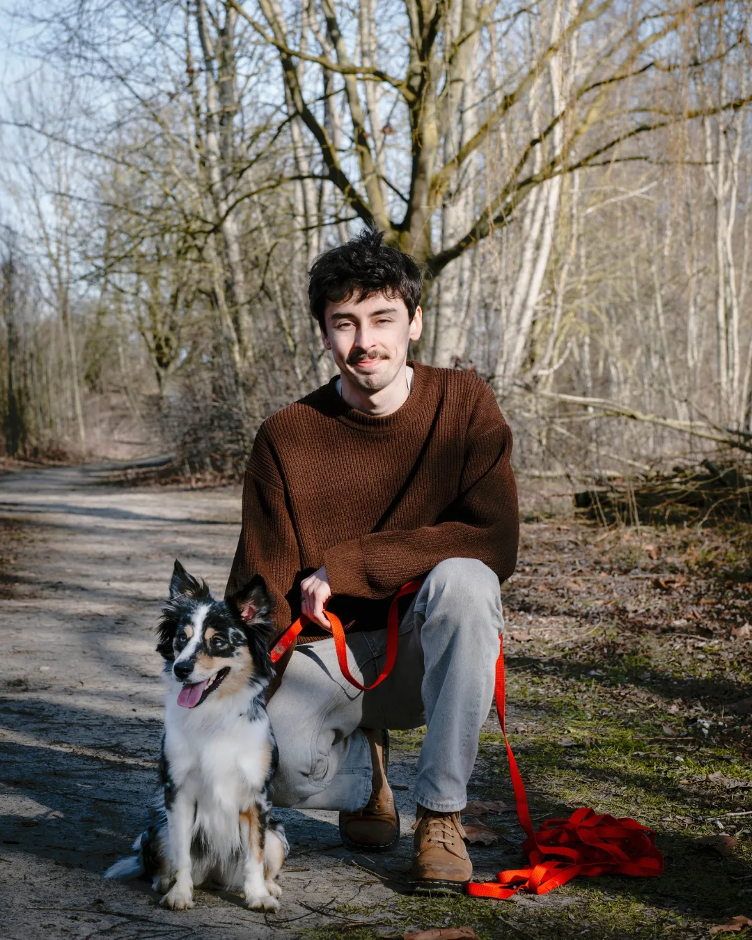 Jonge man met bruin truitje en grijze broek knielt op een bospaadje naast een zittende, zwart-witte hond met rode riem, omringd door kale bomen.