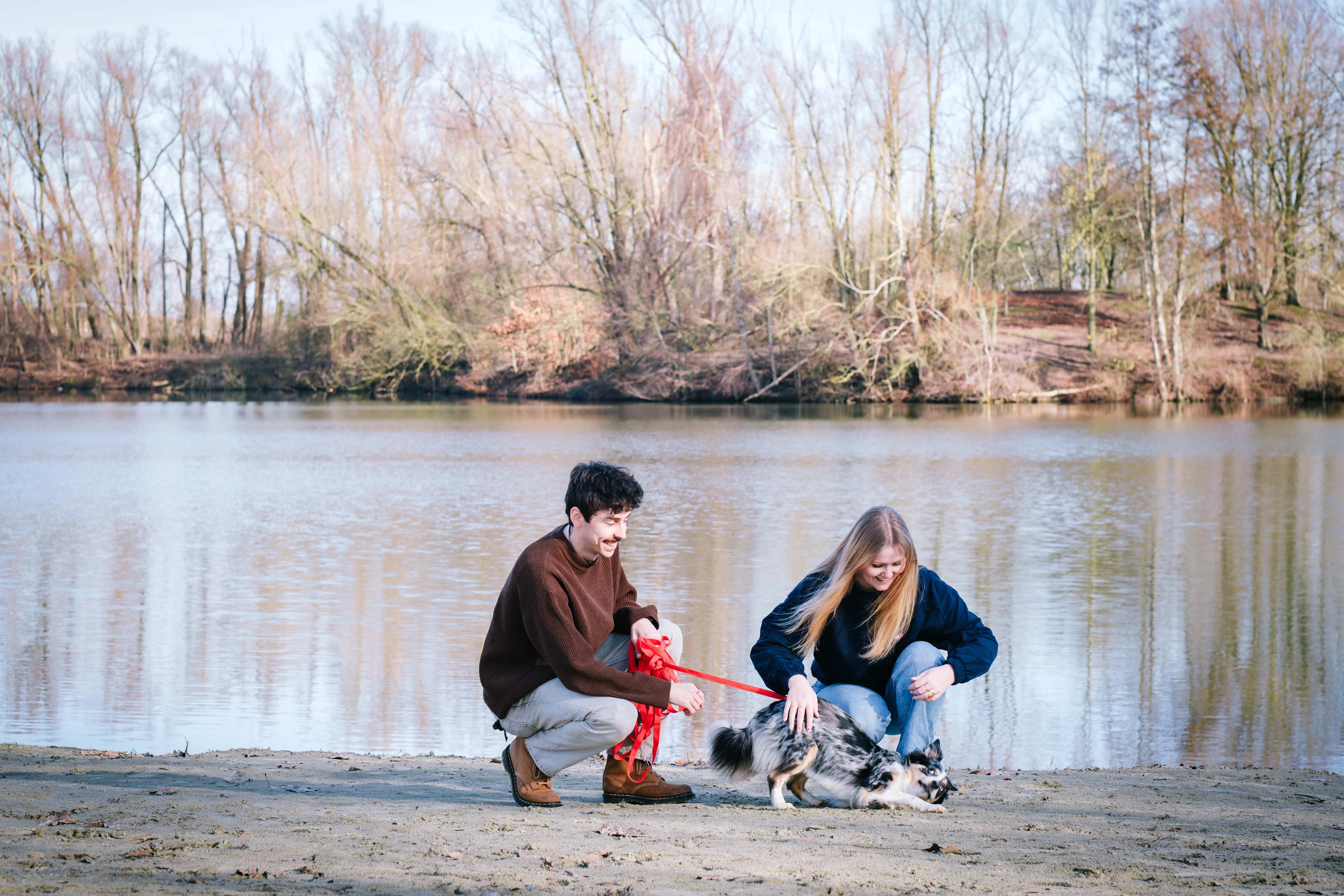 Twee personen hurken bij het water aan een meer met herfstige bomen op de achtergrond; ze spelen met een zwarte-witte hond aan een rode hondenriem op het zand.