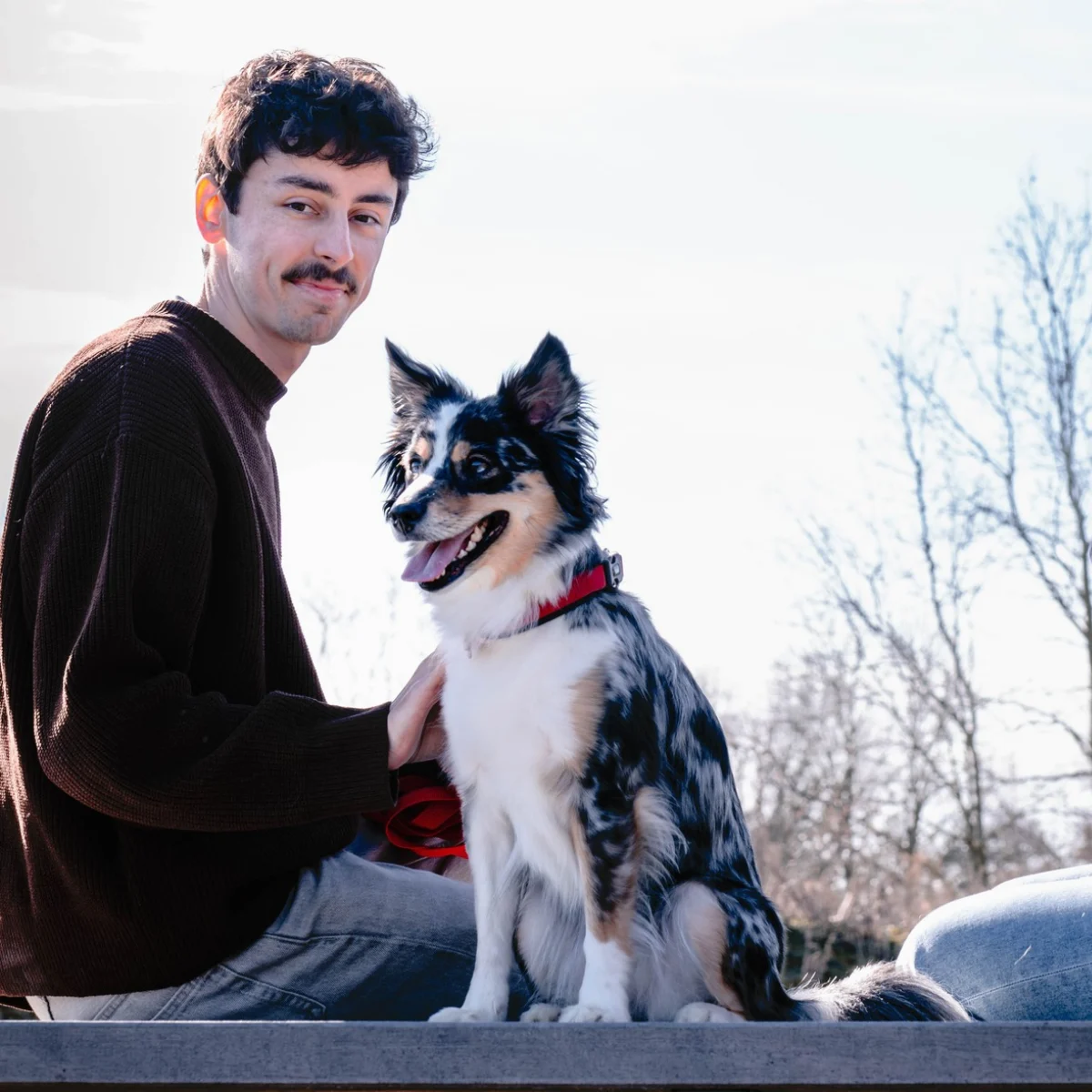 Een man met een snor en een donkere trui zit naast een zwart-witte hond met een rood halsband, buiten met kale bomen op de achtergrond.