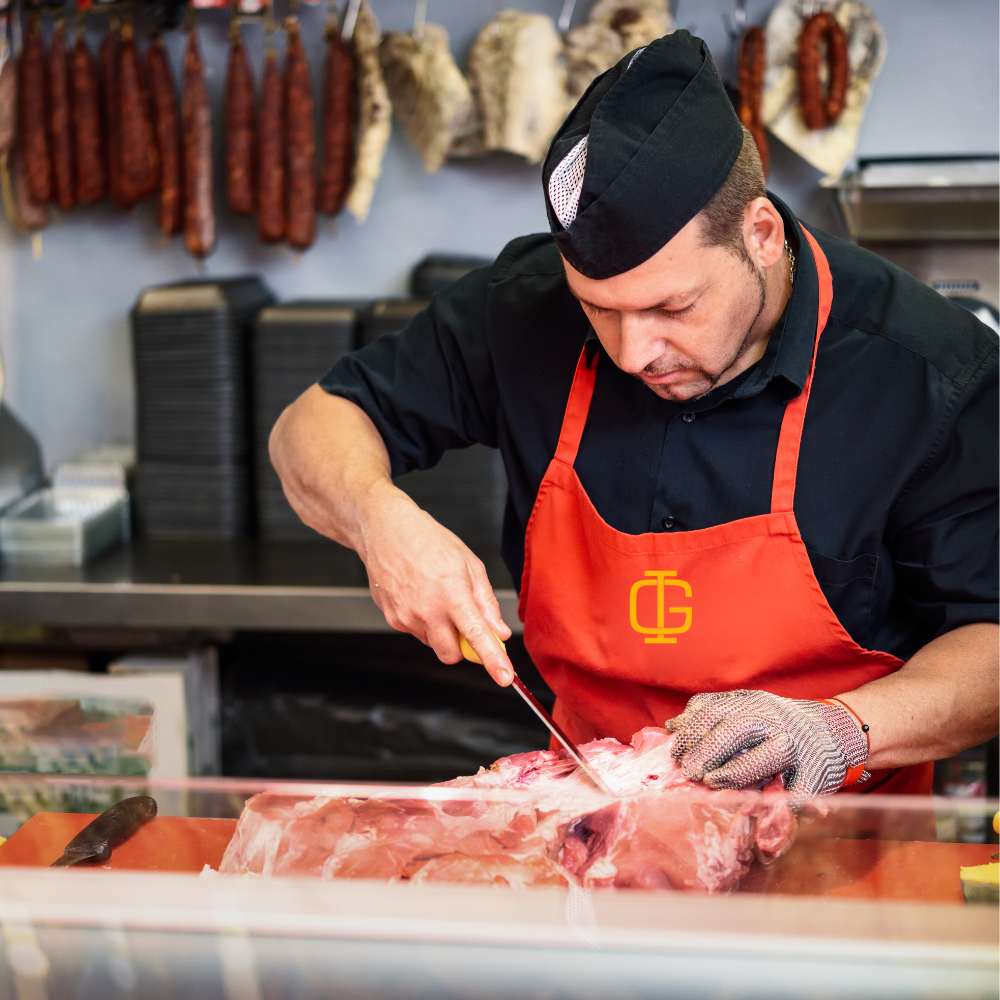 Un boucher portant un tablier rouge et un chapeau noir coupe un morceau de viande dans une boucherie avec des saucisses suspendues en arrière-plan
