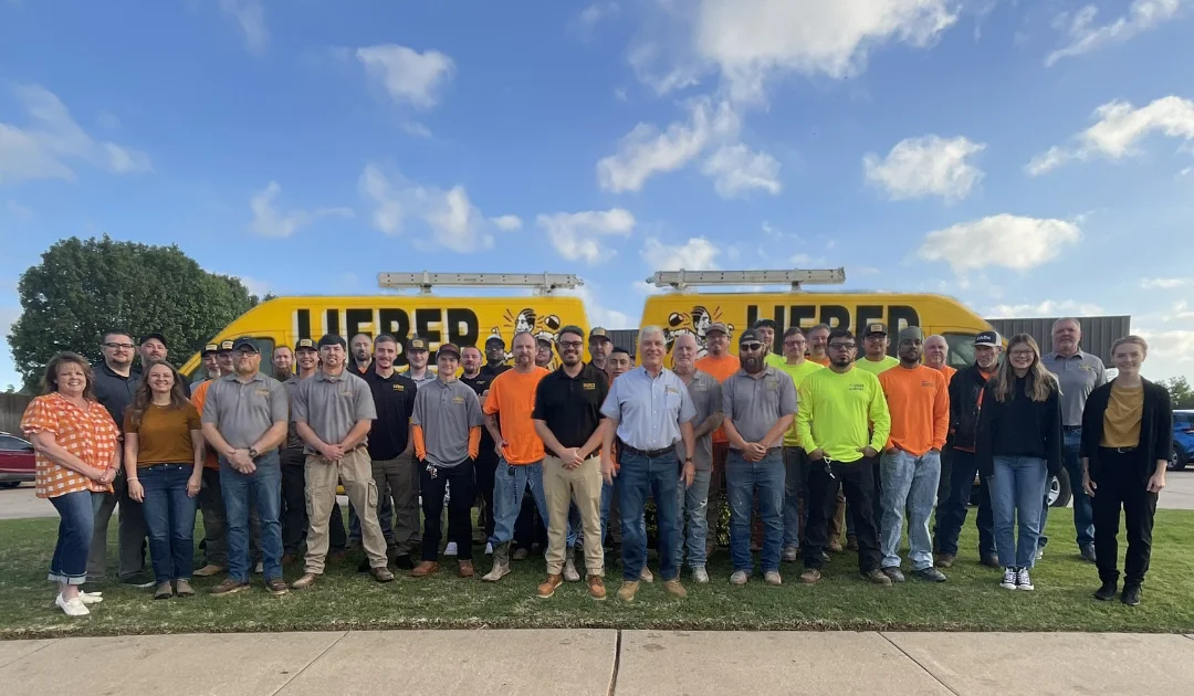 Group photo of Lieber Mechanical Plumbing Heating Air Conditioning team standing outdoors in front of two yellow company vans under a partly cloudy sky.