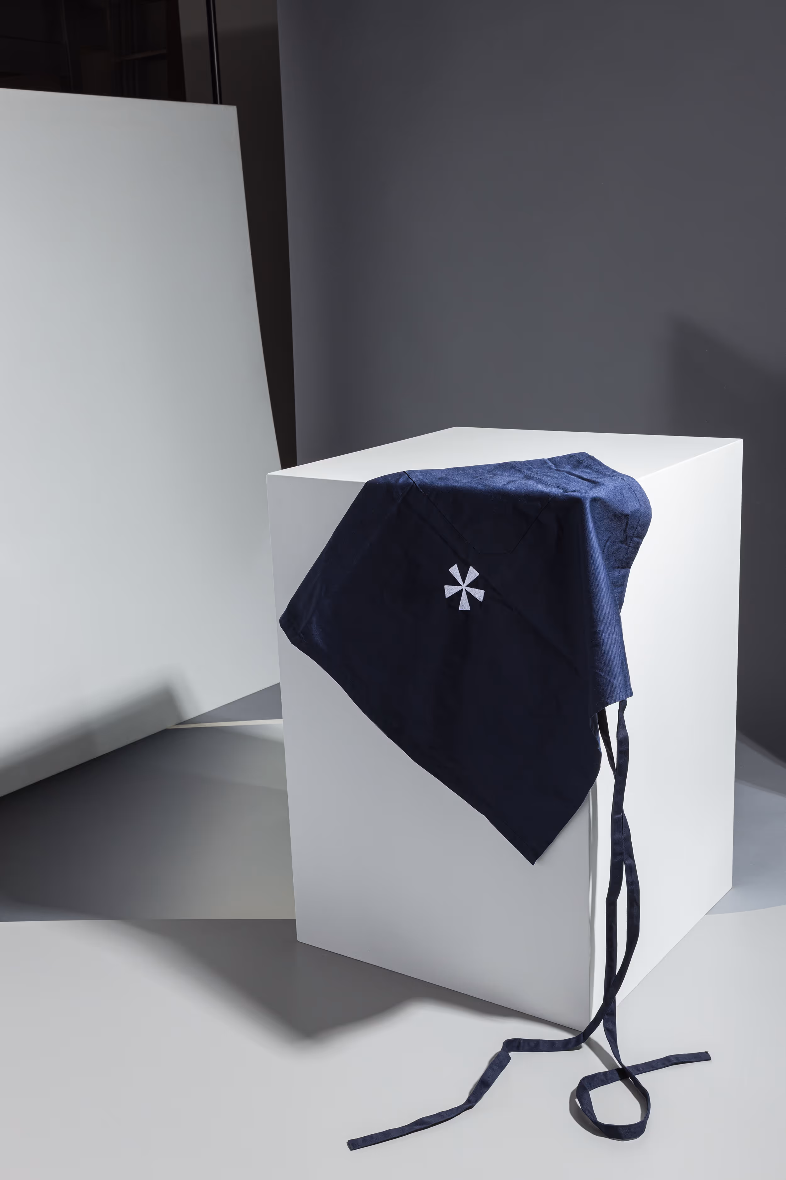Dunkelblaue Arbeitsschürze mit aufgenähtem, weissem Sternsymbol (Asterisk) für das Geigenbauatelier Schutter Widmer, liegend auf weissem Kubus