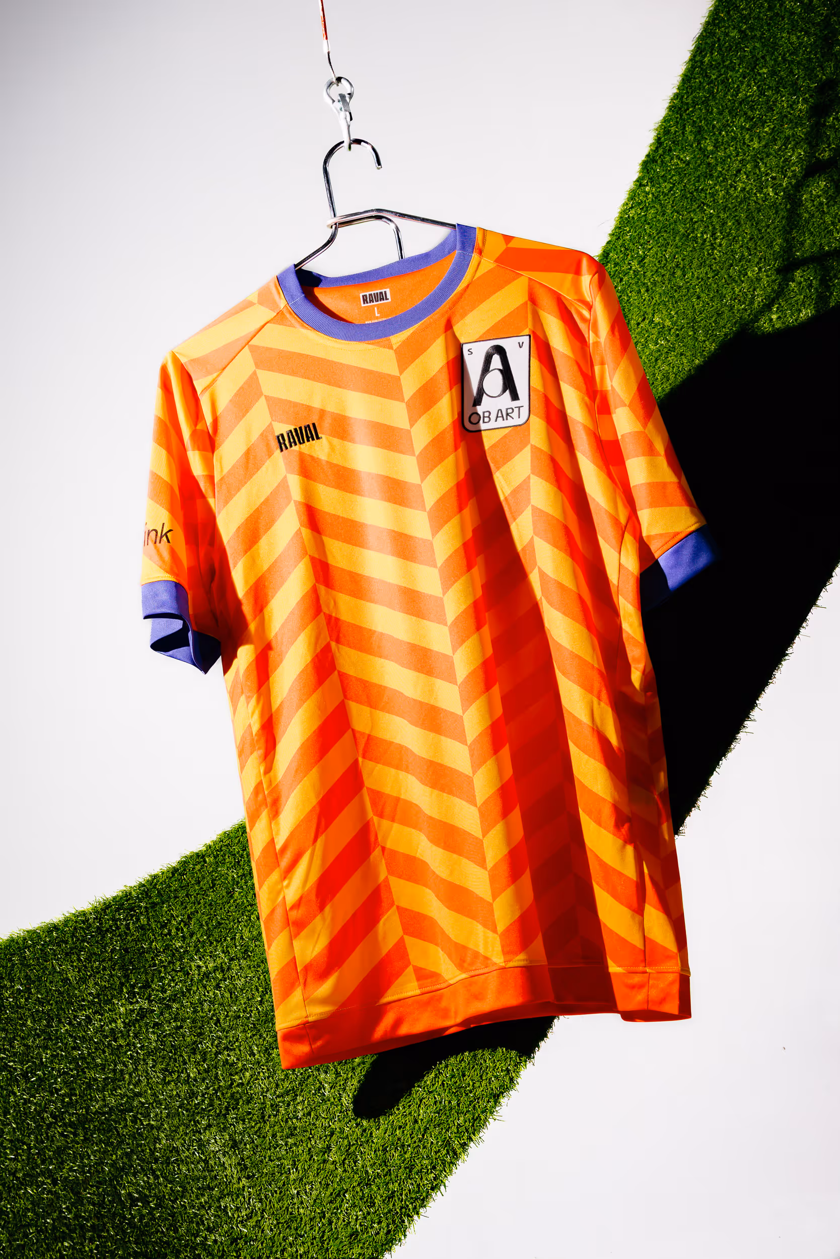 Vorderseite des Orange-Gelb-farbigen Heimtrikot des ObArt Sportvereins, hängend vor einem Stück grünen Kunstrasen