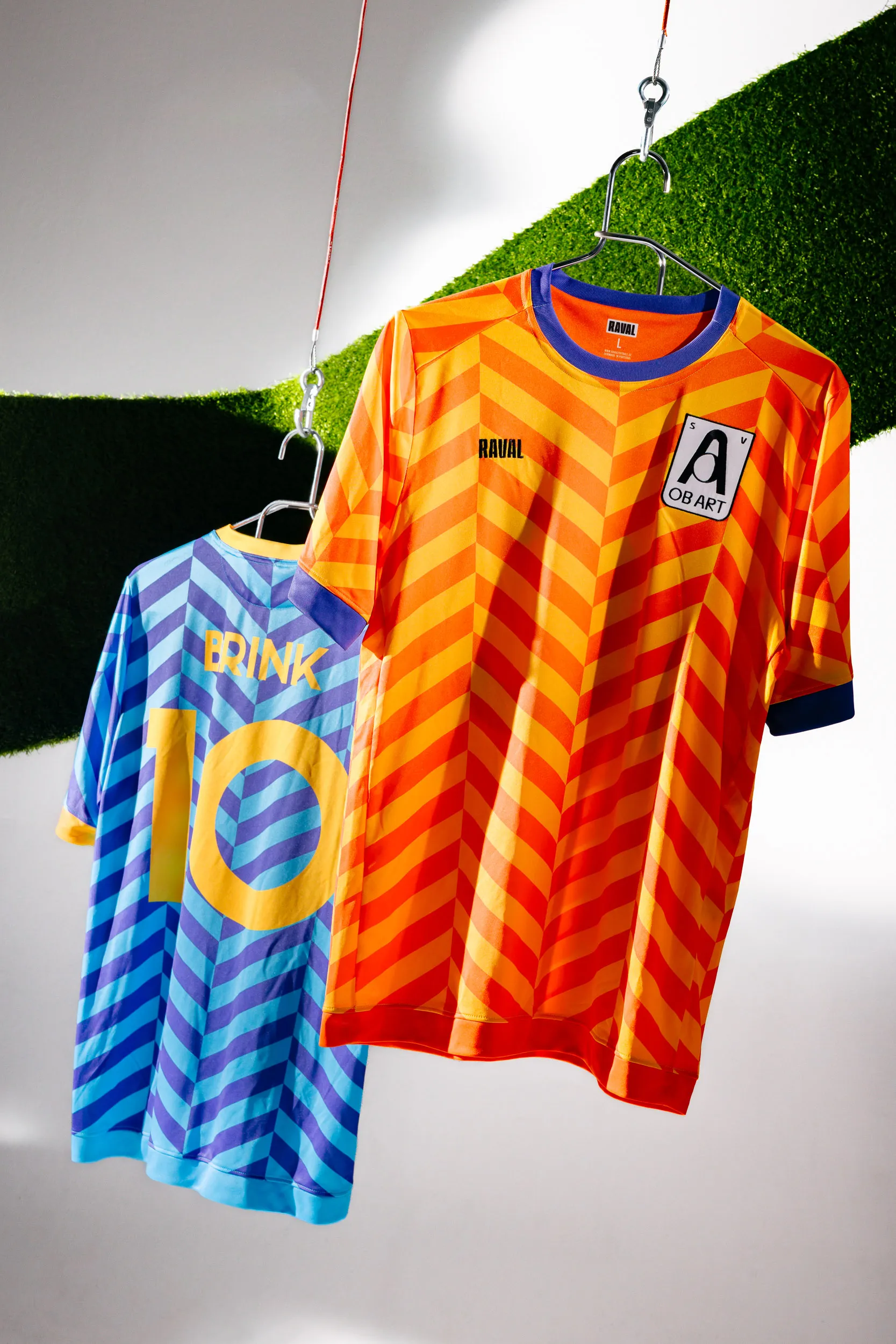 Orange-Gelb-farbiges Heimtrikot und Blau-Violett-farbiges Auswärtstrikot des ObArt Sportvereins, hängend vor einem Stück grünen Kunstrasen