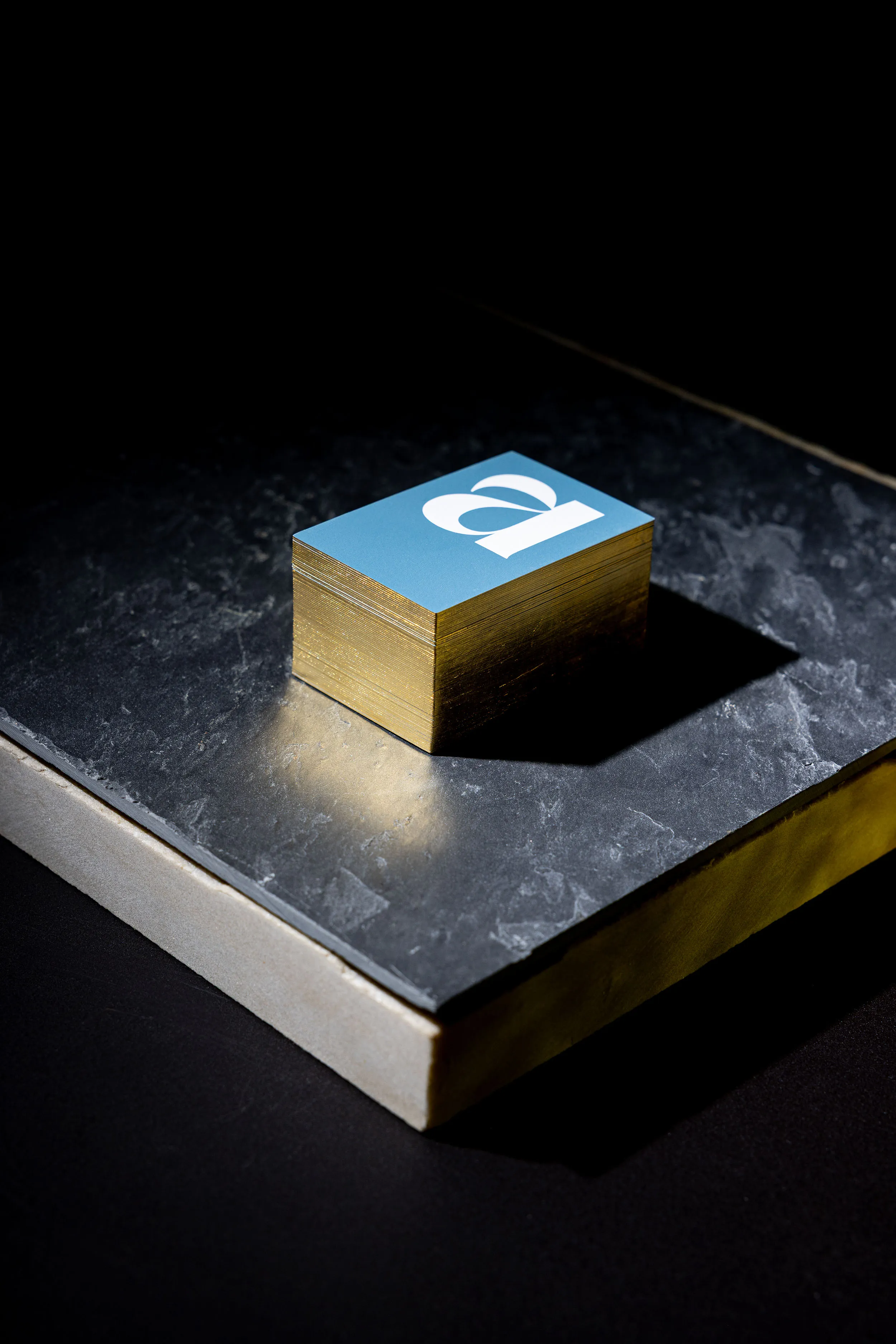 Stapel von Visitenkarten mit blauem Papier und Gold reflektierendem Farbschnitt für das Goldschmiedatelier arany, stehend auf dicker Steinplatte, vor dunklem Hintergrund