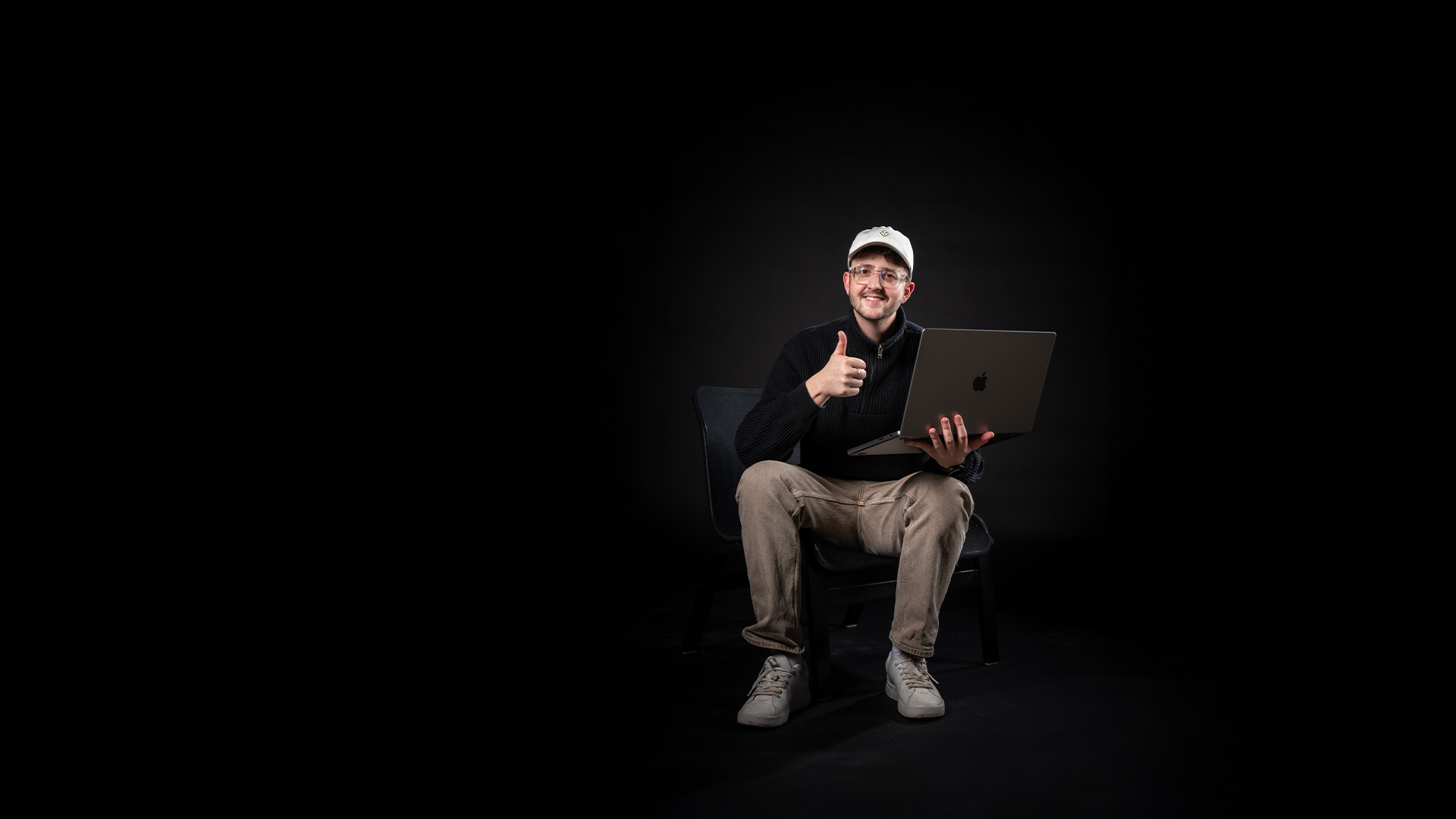Luca David, Content Creator und Social Media Experte, sitzt mit einem Laptop auf dem Schoss auf einem Stuhl vor schwarzem Hintergrund und zeigt Daumen hoch – moderner Arbeitsplatz für digitales Storytelling.