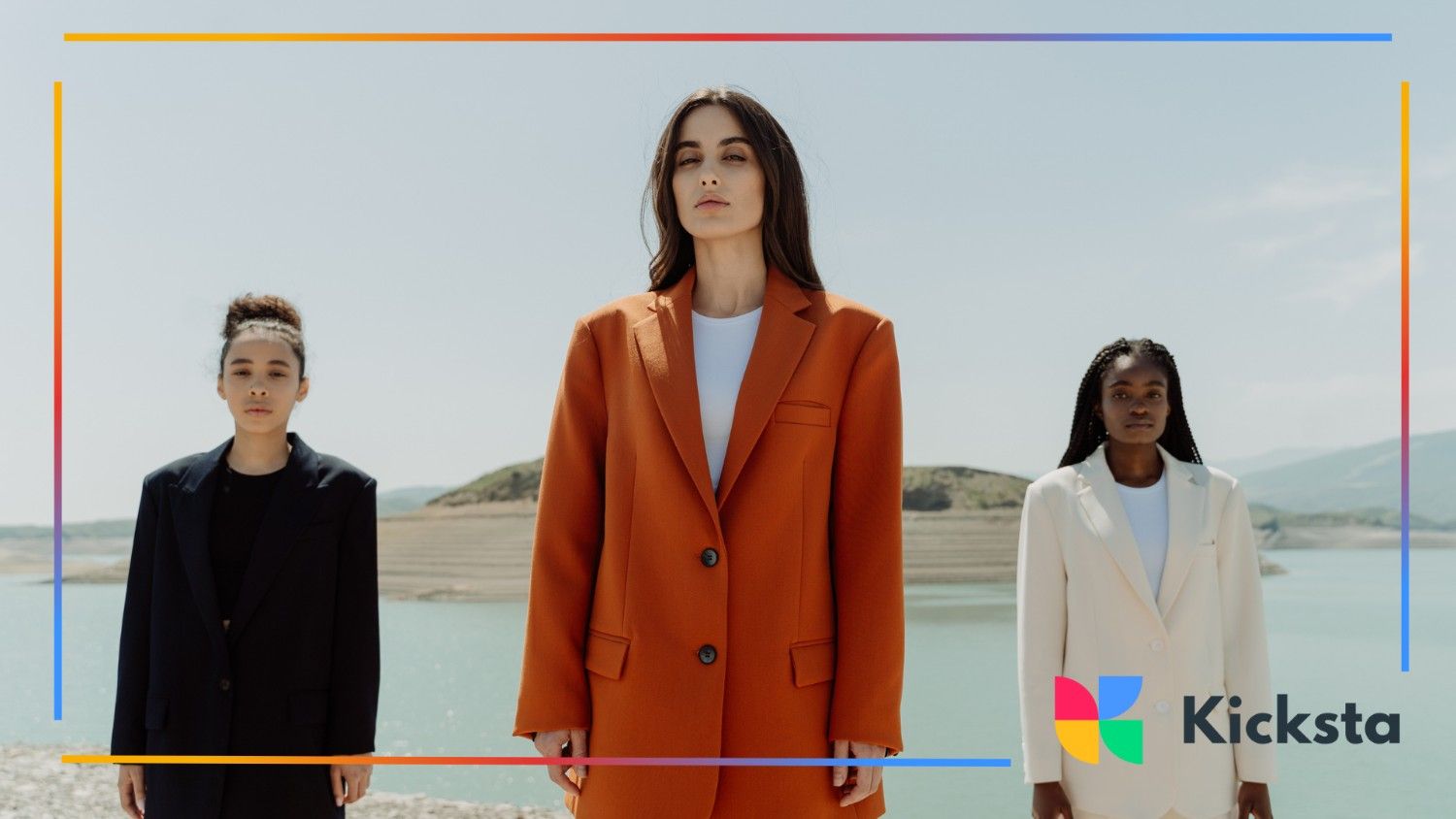 Three women in bold blazers (orange, white, and navy) standing confidently in front of a scenic lake and mountain background