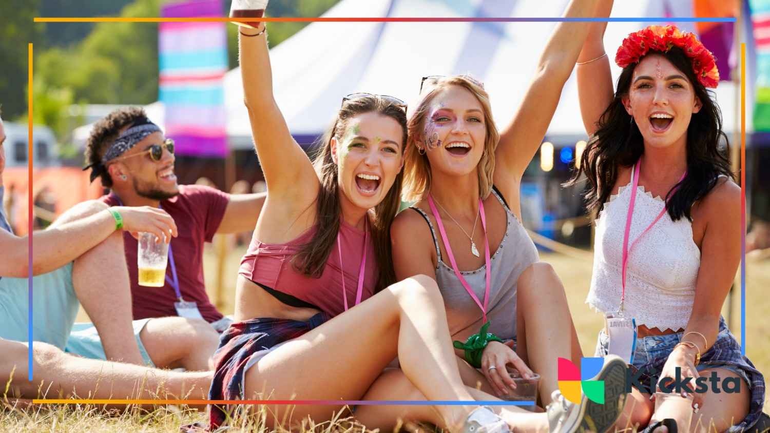 Group of friends sitting on grass at a music festival, smiling and cheering with face paint and drinks in hand