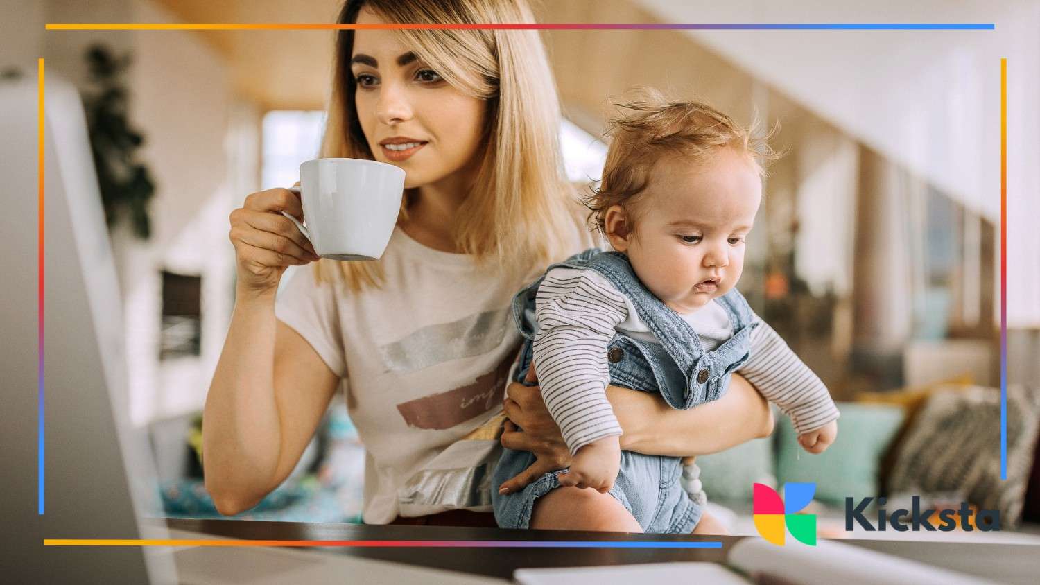 A mom multitasks—sipping coffee, holding her baby, and working on a laptop at home