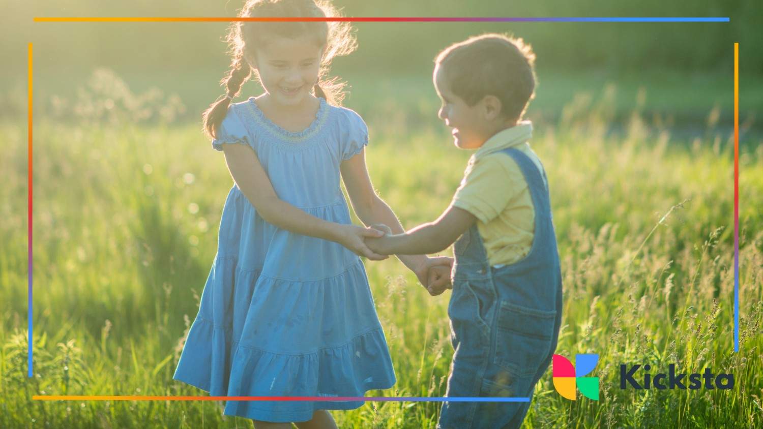 A girl in a blue dress and a boy in overalls hold hands and smile in a grassy meadow