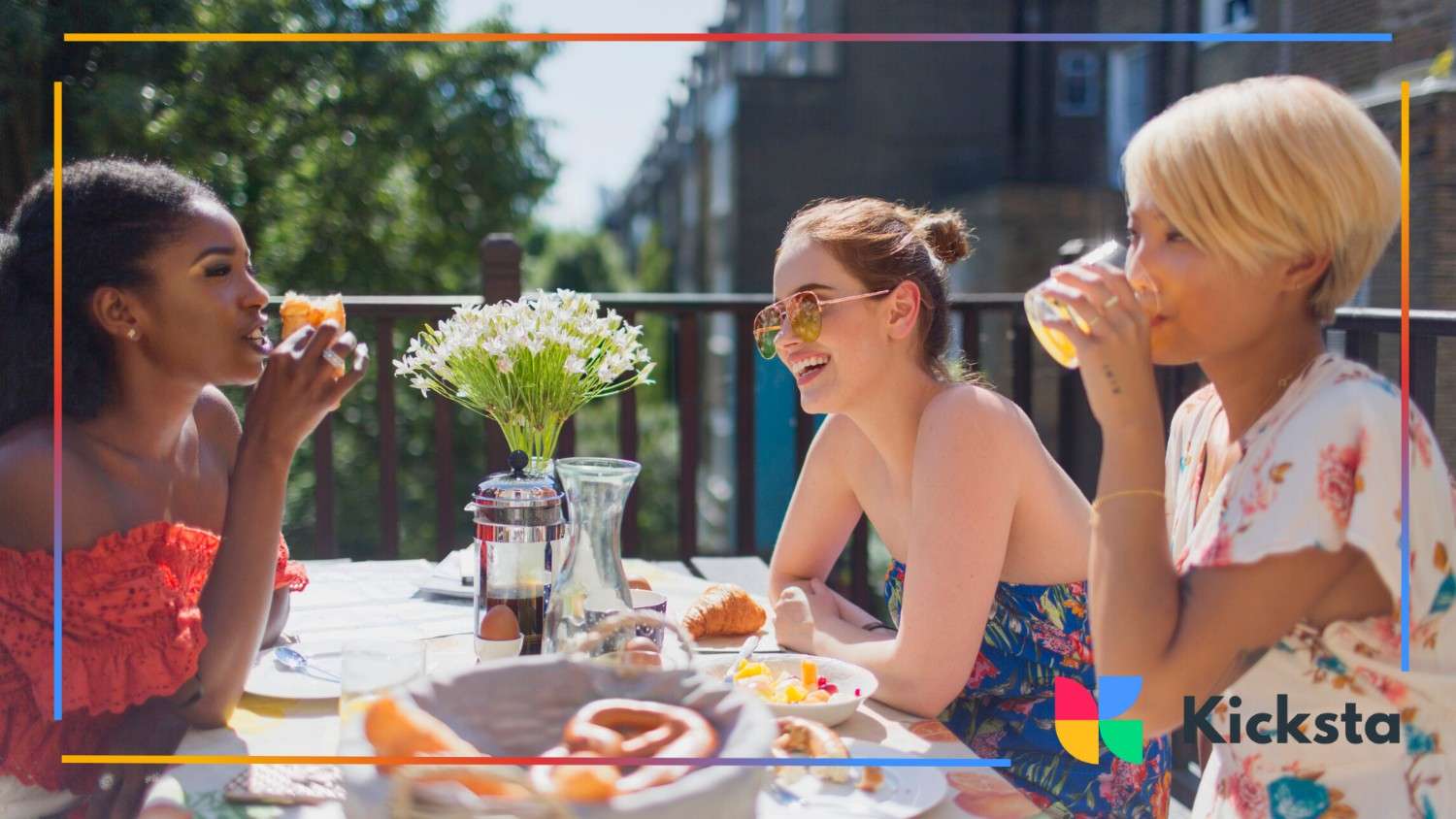 Three stylish women enjoying a sunny brunch on a balcony, surrounded by fresh food and laughter