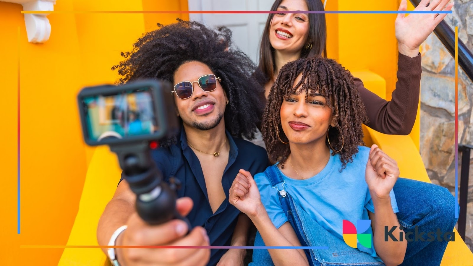 Three friends smiling and posing for a video selfie with a camera on a stick in front of a colorful wall.