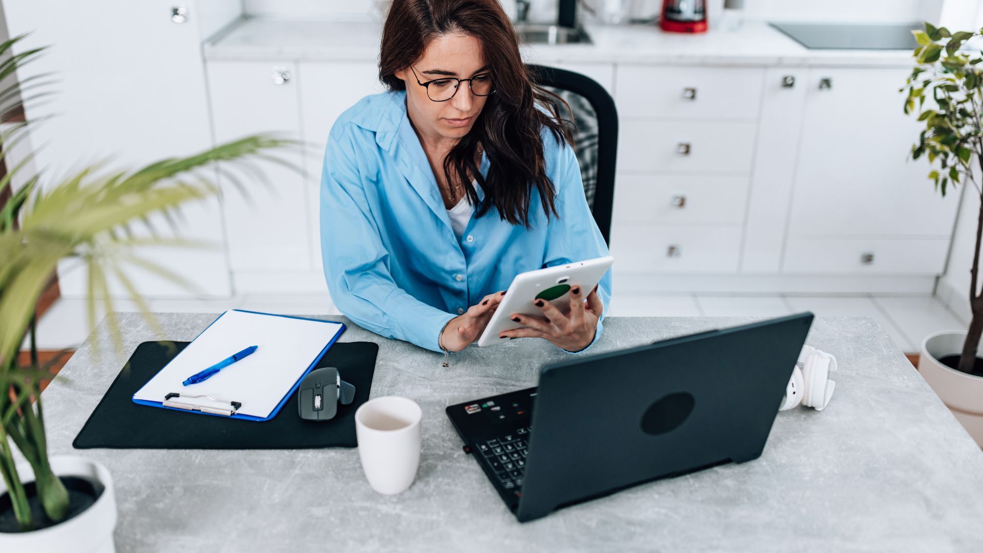 Woman working from home, sitting at a kitchen table with a laptop and tablet, reviewing notes with a clipboard nearby.