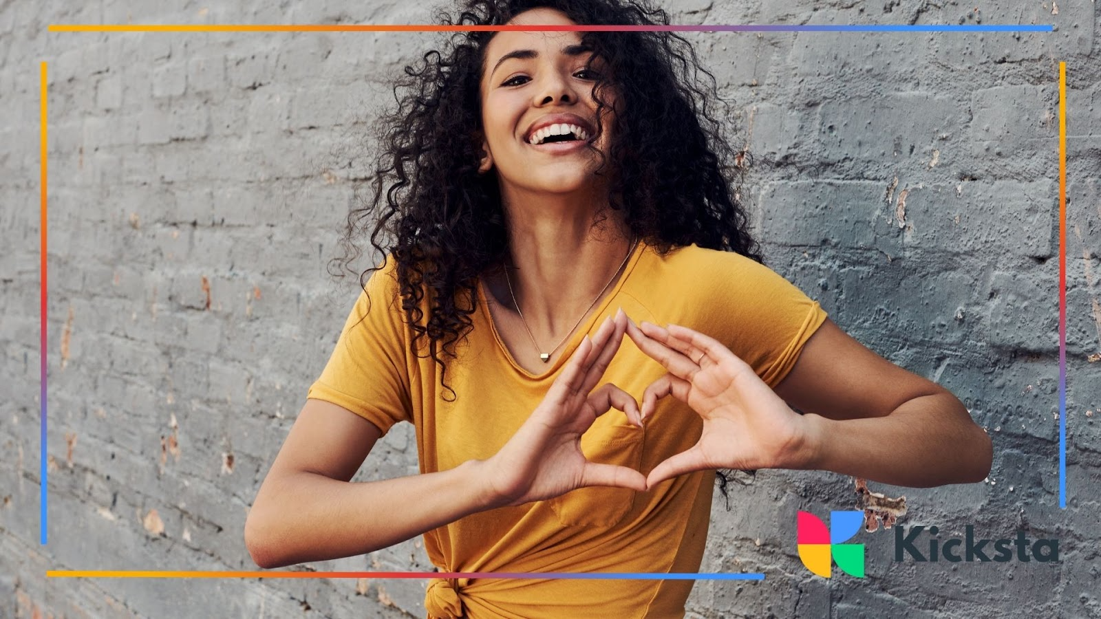 A curly-haired woman smiles big while forming a heart with her hands