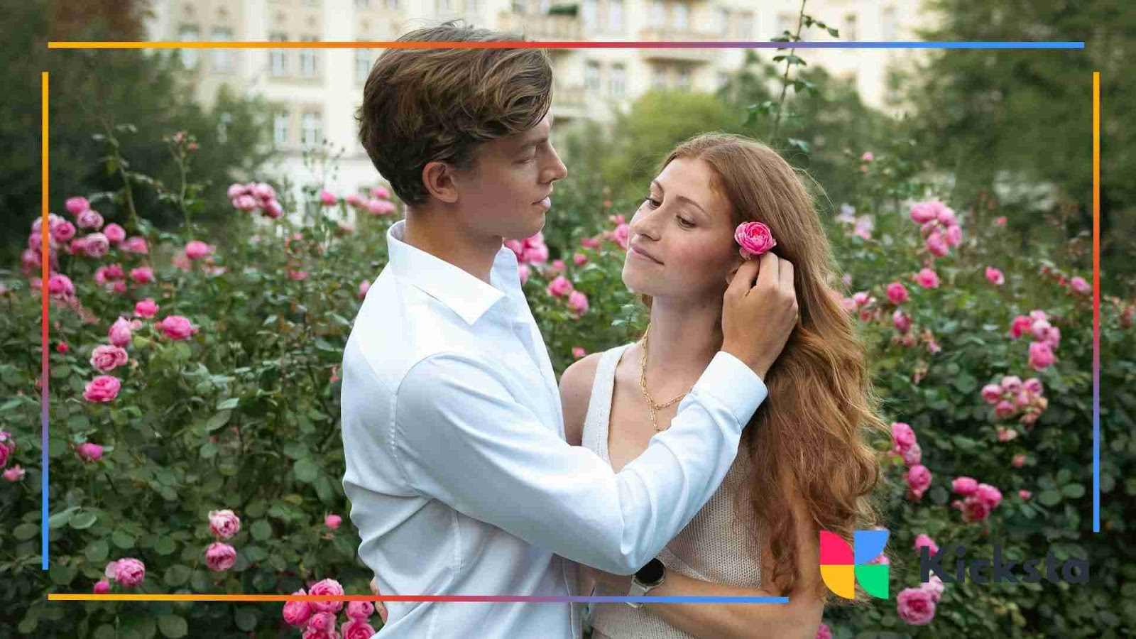 A young man placing a pink rose behind a woman’s ear as they stand surrounded by blooming rose bushes