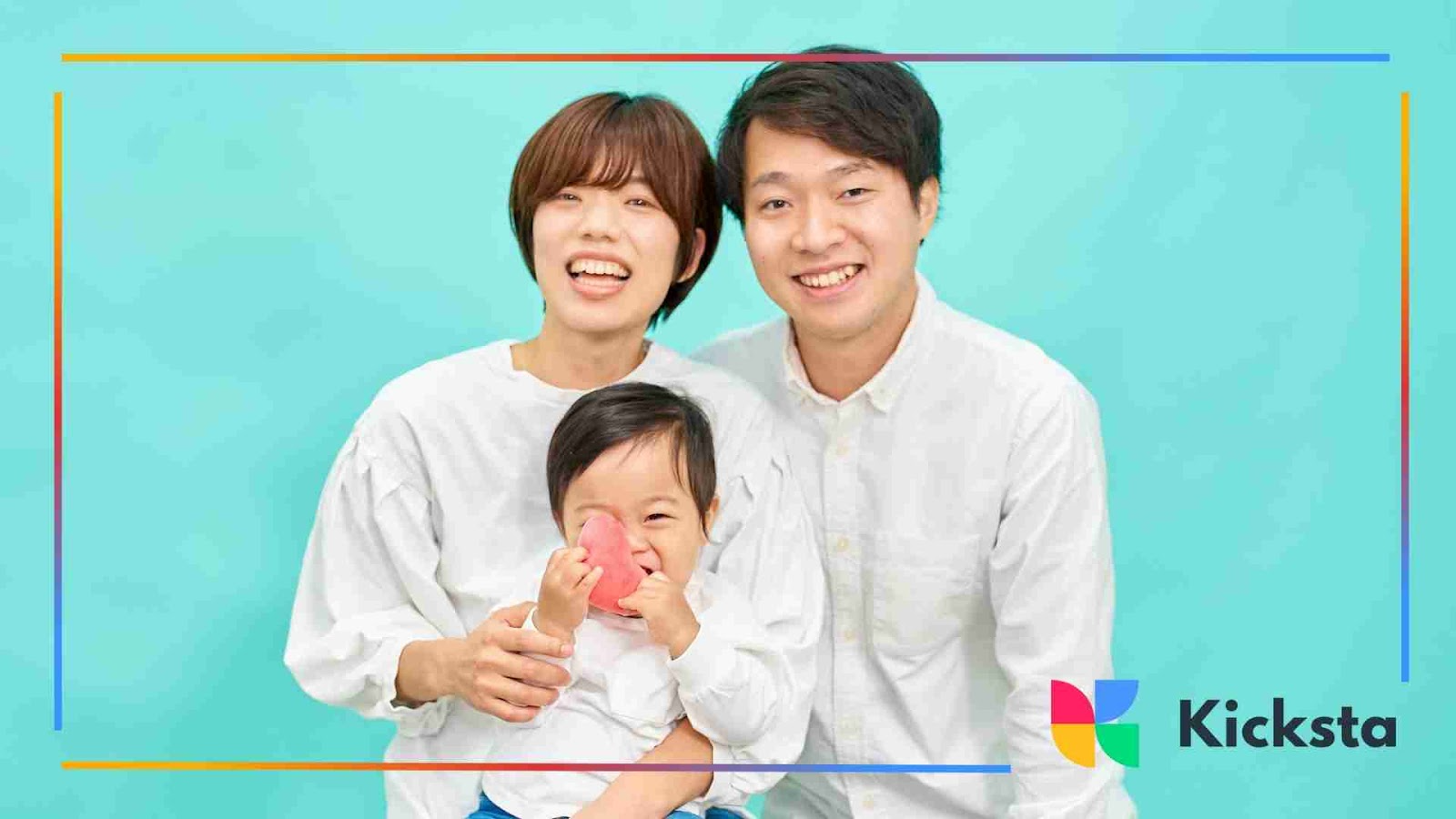 A smiling family of three posing in front of a turquoise background, with the baby holding a red heart-shaped toy