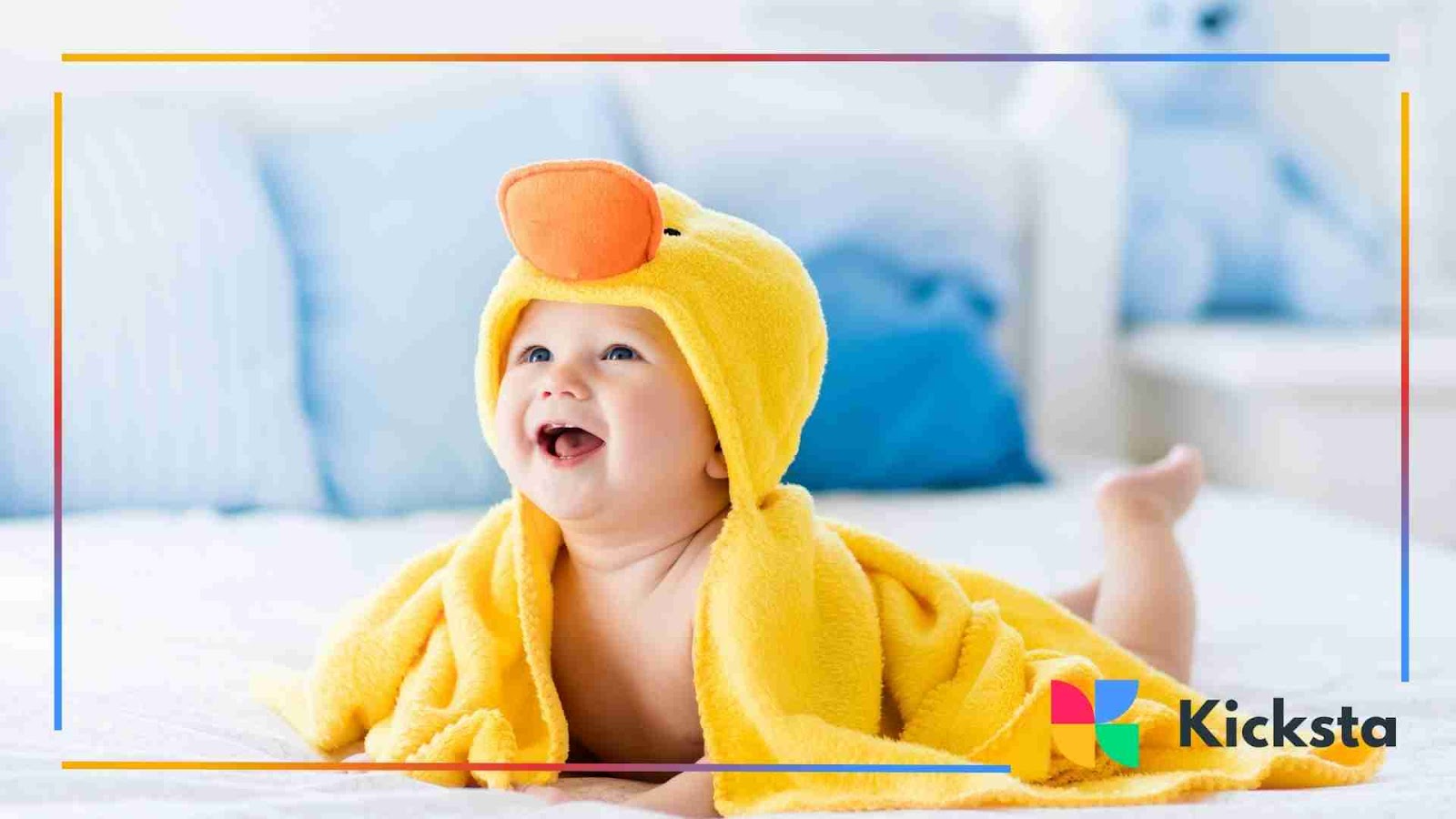 A baby wrapped in a bright yellow towel with a duck hood, lying on a bed and laughing