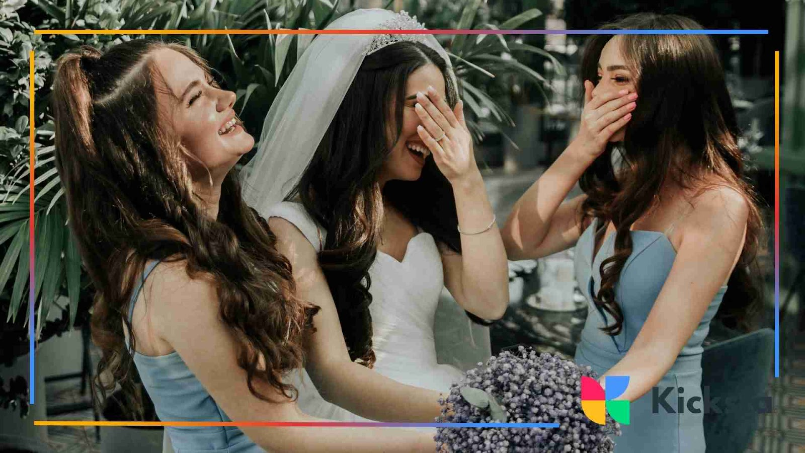 A bride and two bridesmaids laughing together, dressed in wedding attire surrounded by greenery