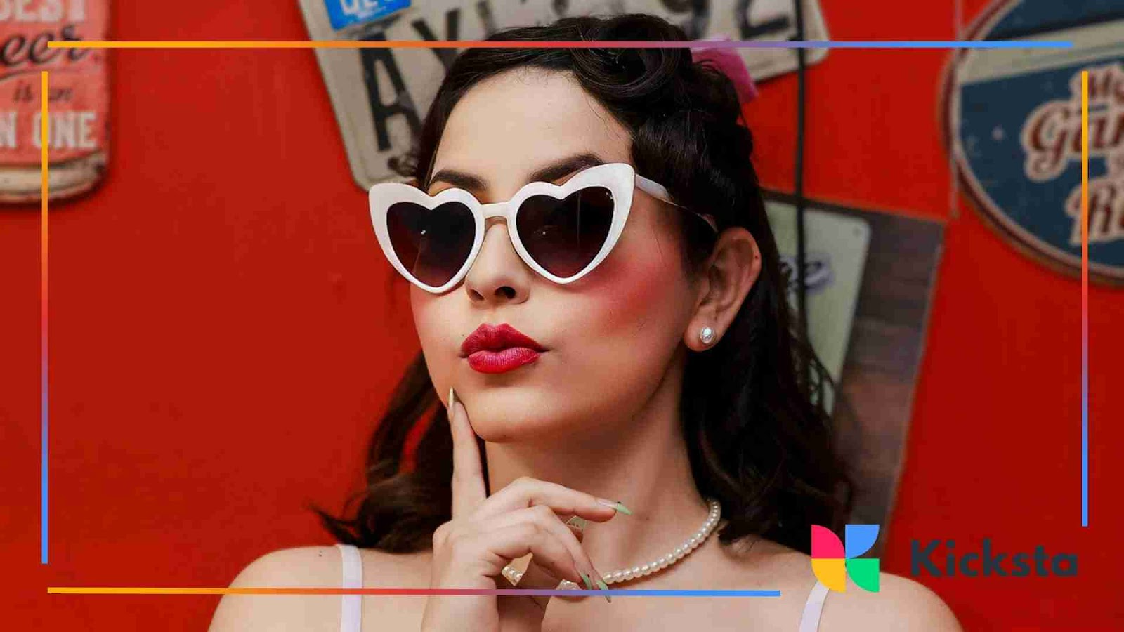 Woman with short dark hair wearing white heart-shaped sunglasses, red lipstick, and pearl necklace posing against a red wall