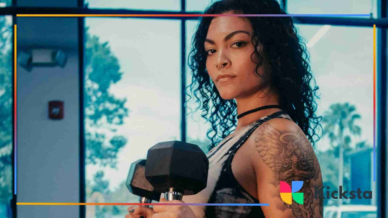 Woman with curly hair and tattoos holding dumbbells in a gym setting