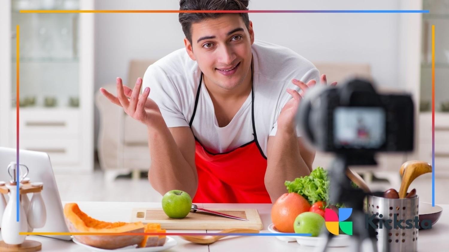 Man wearing a red apron recording a cooking video in the kitchen, surrounded by fruits and vegetables