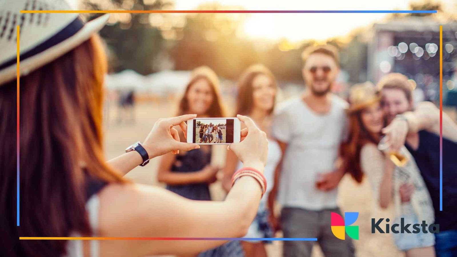 Person wearing a hat taking a smartphone photo of friends smiling together at an outdoor festival during sunset