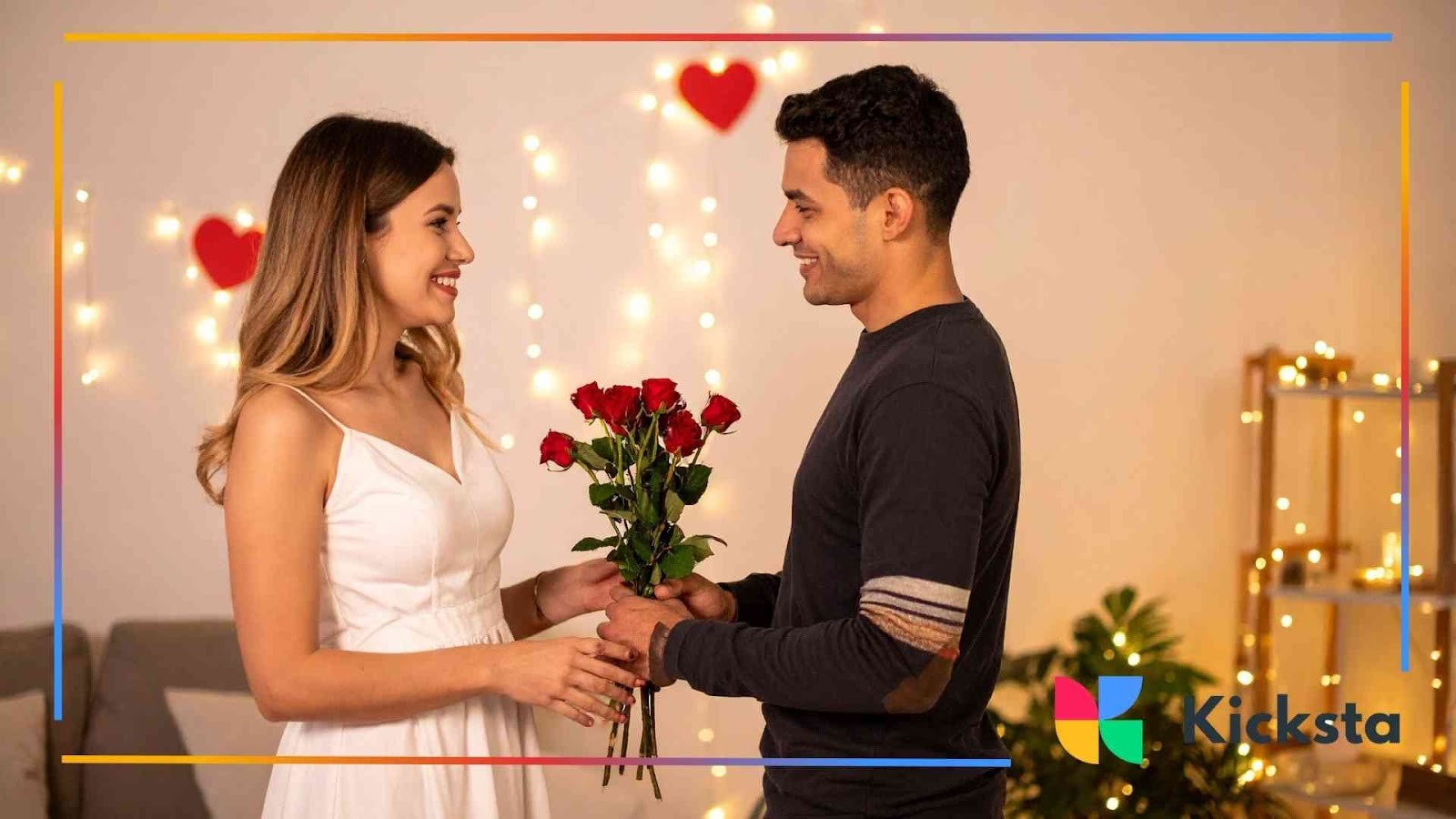 Smiling man giving a bouquet of red roses to a woman in a white dress with heart decorations in the background.
