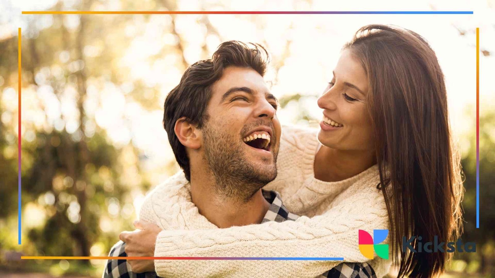 Happy couple laughing outdoors in warm sunlight, woman hugging the man from behind.