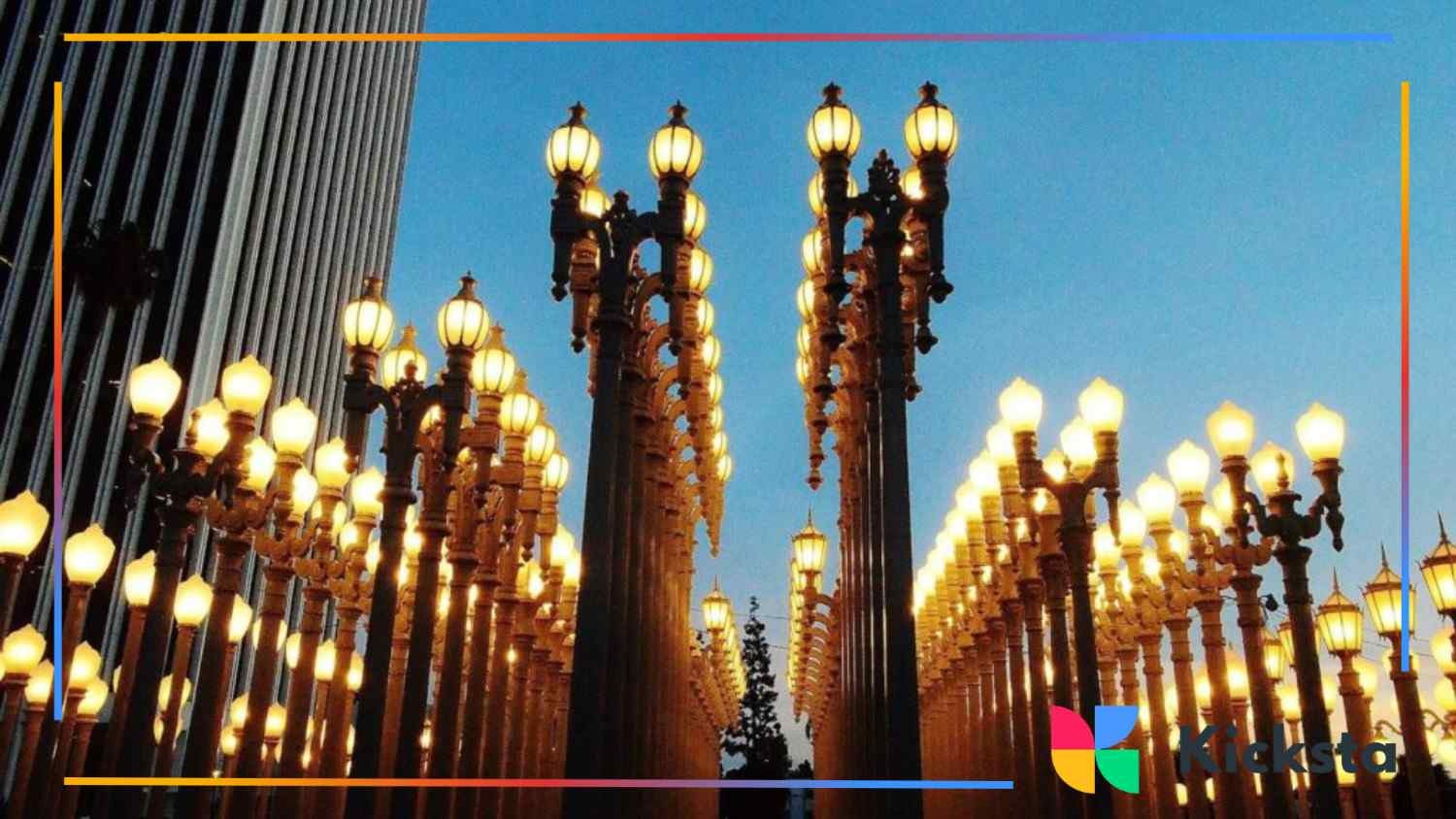 Rows of tall, glowing street lamps lined up between city buildings at dusk.