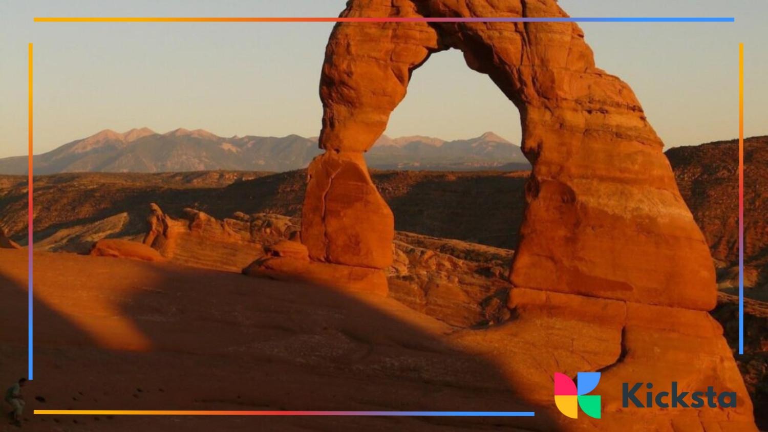 Iconic Delicate Arch rock formation in Arches National Park glowing in warm sunset light against a mountain backdrop.