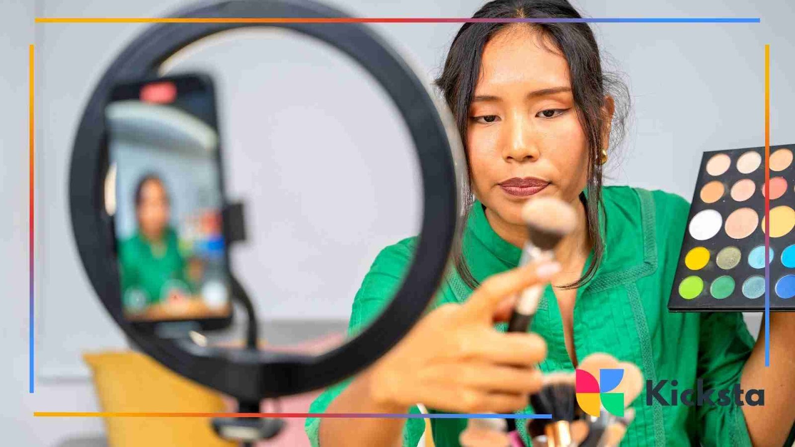 A woman wearing a green shirt filming a makeup tutorial using a smartphone and ring light. She holds a makeup brush and an eyeshadow palette