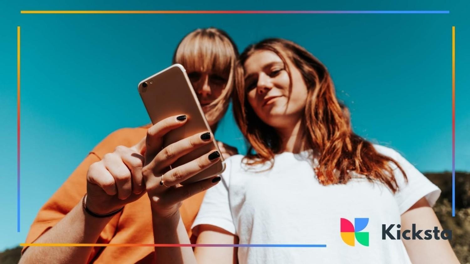 Two young women standing outdoors under a clear blue sky, looking at a smartphone together.