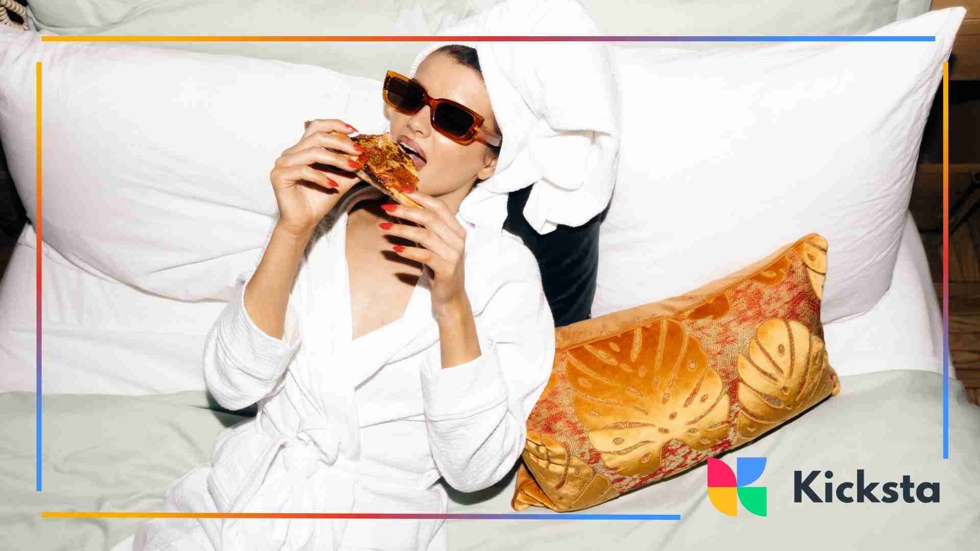 Woman in a bathrobe and sunglasses eating pizza while lounging on a bed.