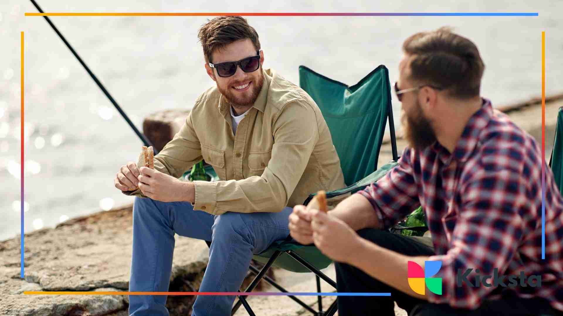 Two men sitting by the water, eating and talking while relaxing in camping chairs.