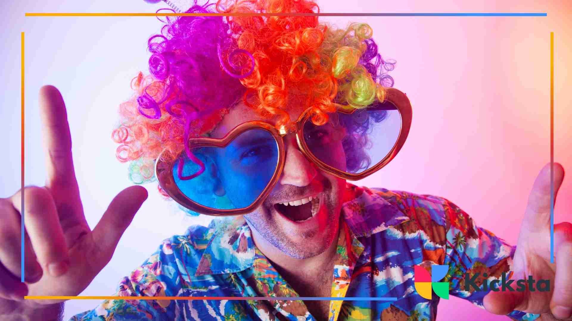 Man wearing a rainbow wig and oversized heart-shaped glasses smiling playfully.