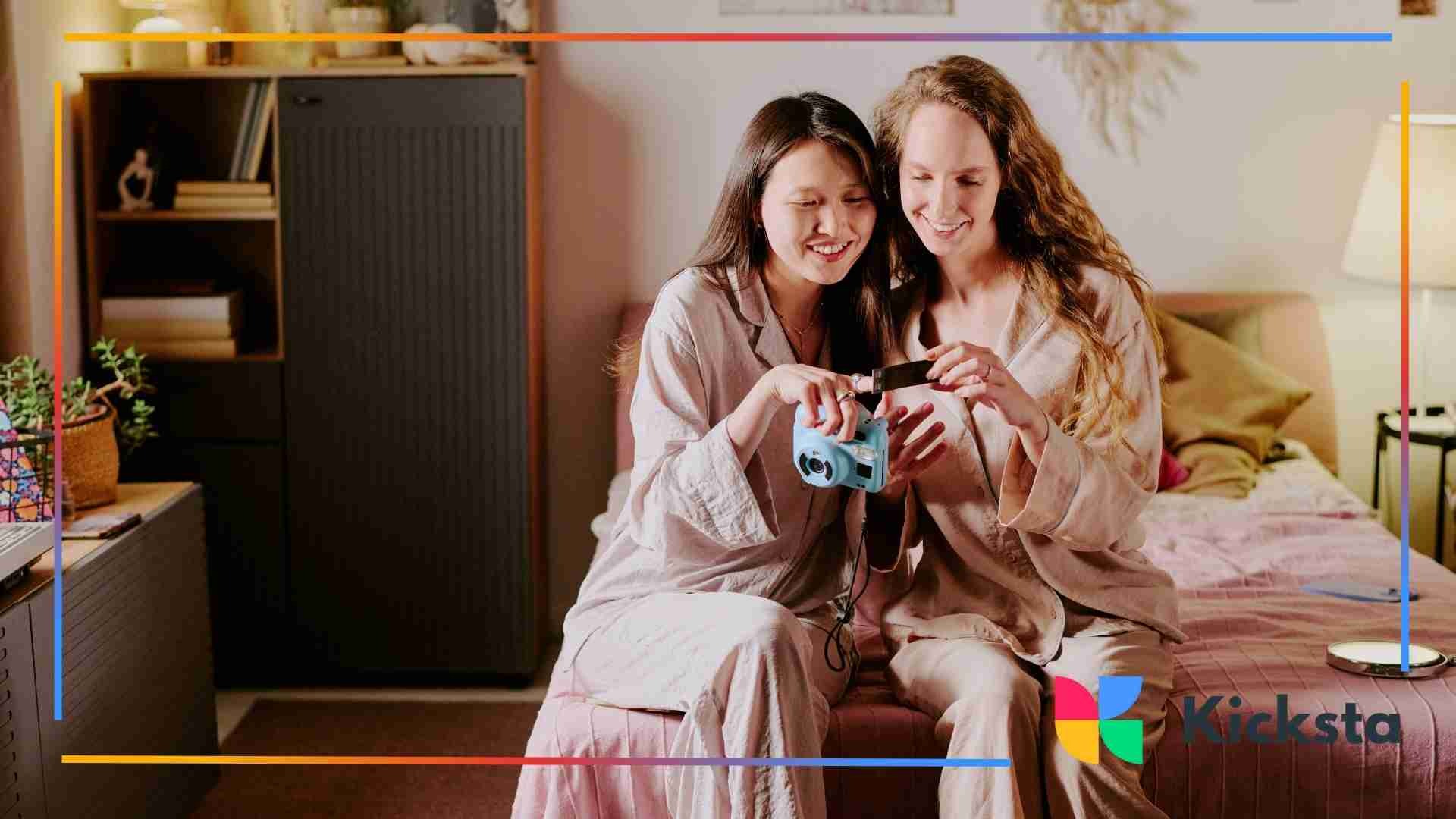 Two women in pajamas sitting on a bed, smiling while looking at a camera together.