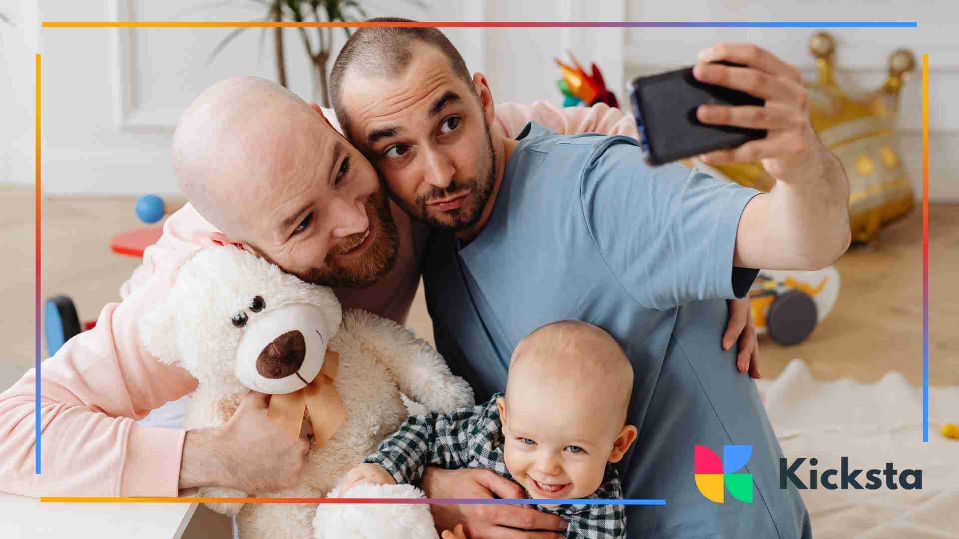 Two men taking a selfie with a baby and a large teddy bear in a brightly lit playroom.