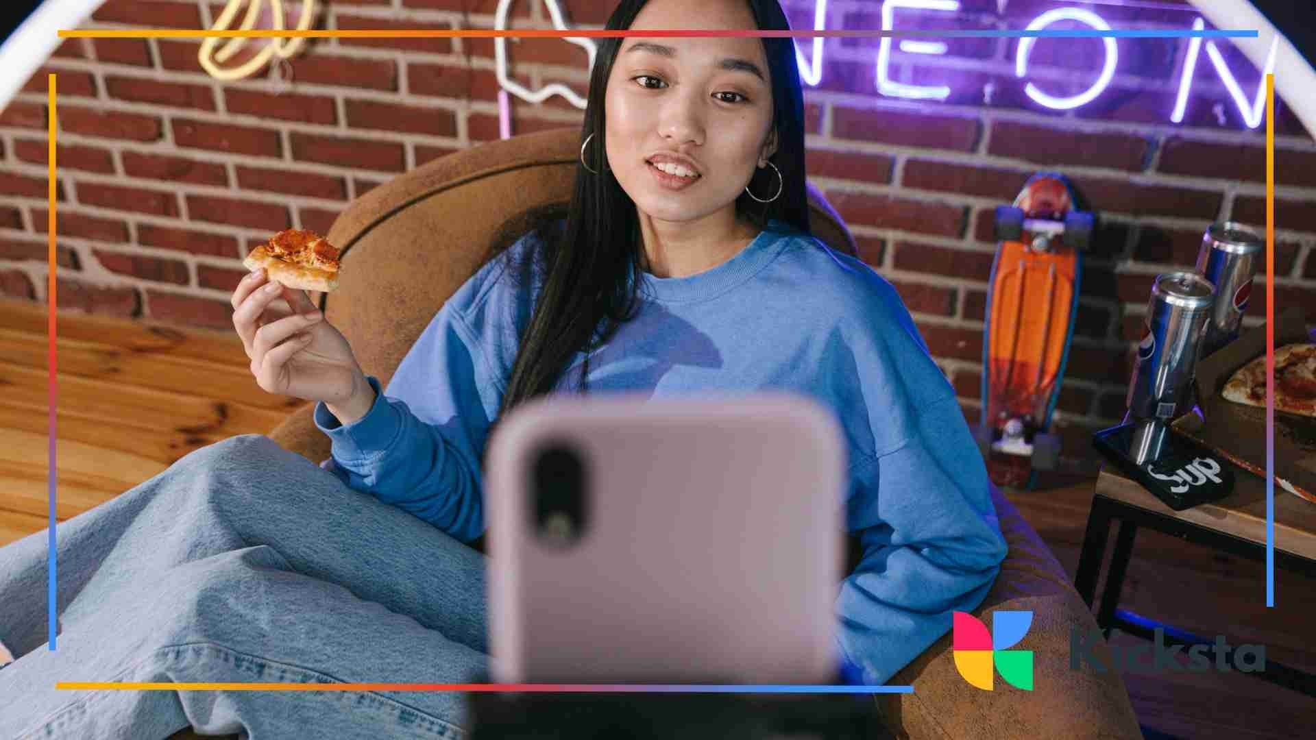 Young woman in a blue sweatshirt recording a video while eating pizza in a cozy room with neon lights and a skateboard in the background.