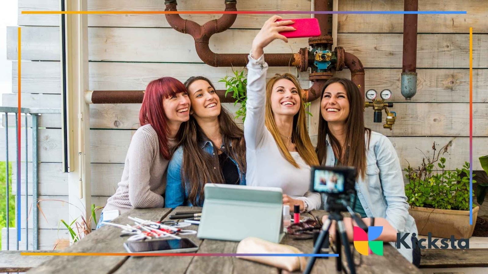 Lifestyle influencers creating group content, smiling for selfie at outdoor café with makeup kits and camera equipment.