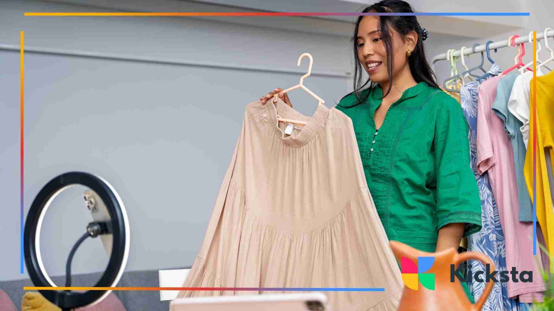 Woman in a green shirt showing a beige dress on camera while filming a fashion video with a ring light and clothes rack in the background.