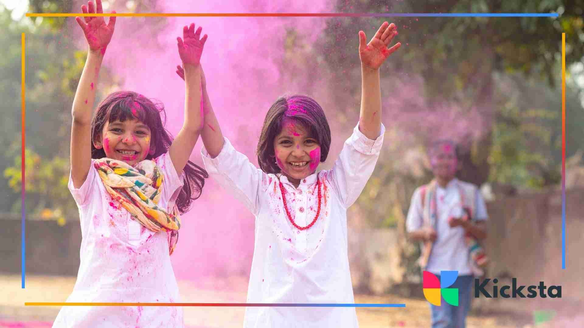 Two young girls covered in pink Holi powder, smiling and raising their hands joyfully outdoors.