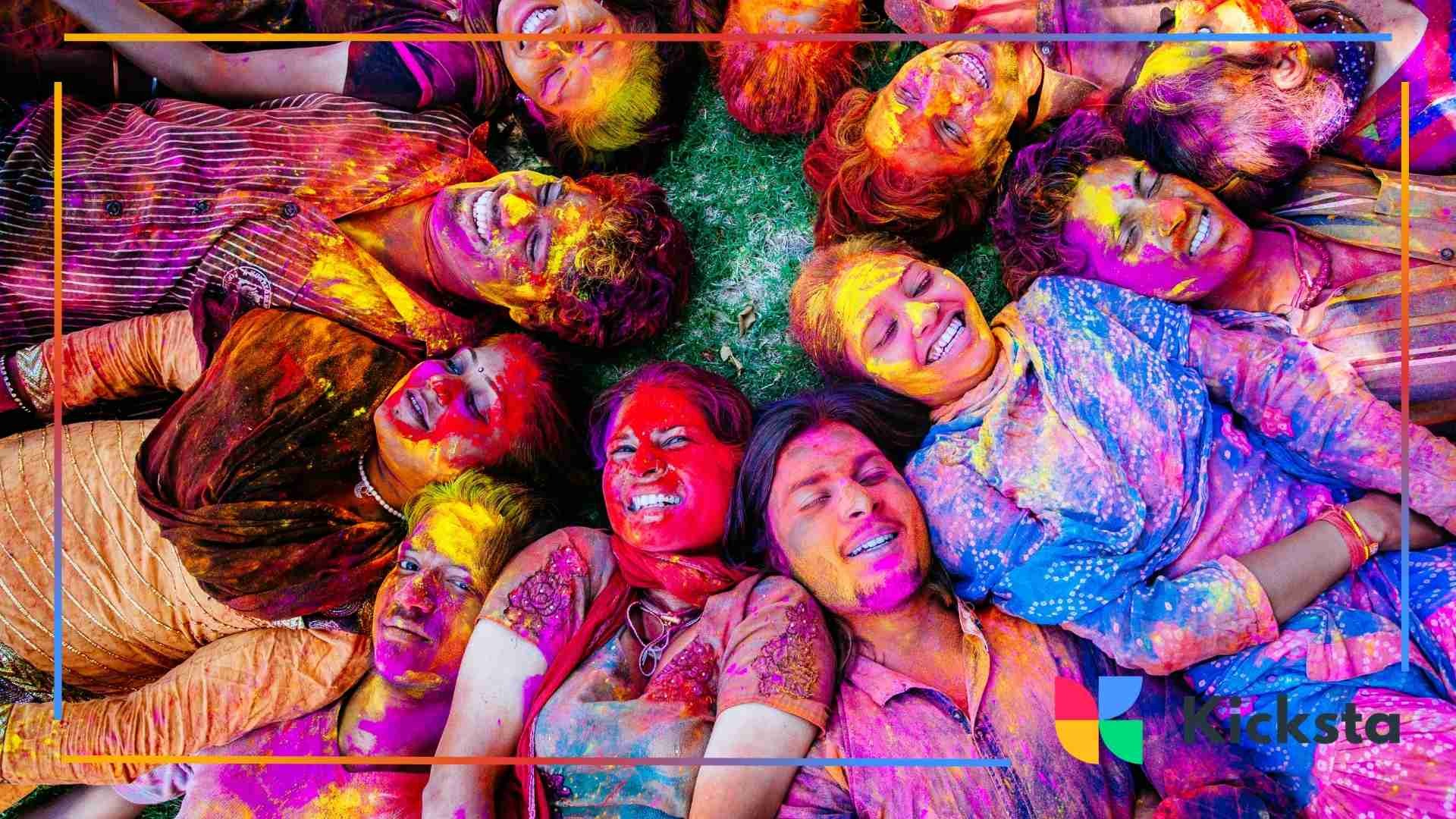 Group of friends lying on the grass in a circle, their faces covered in bright Holi colors, laughing and celebrating together.