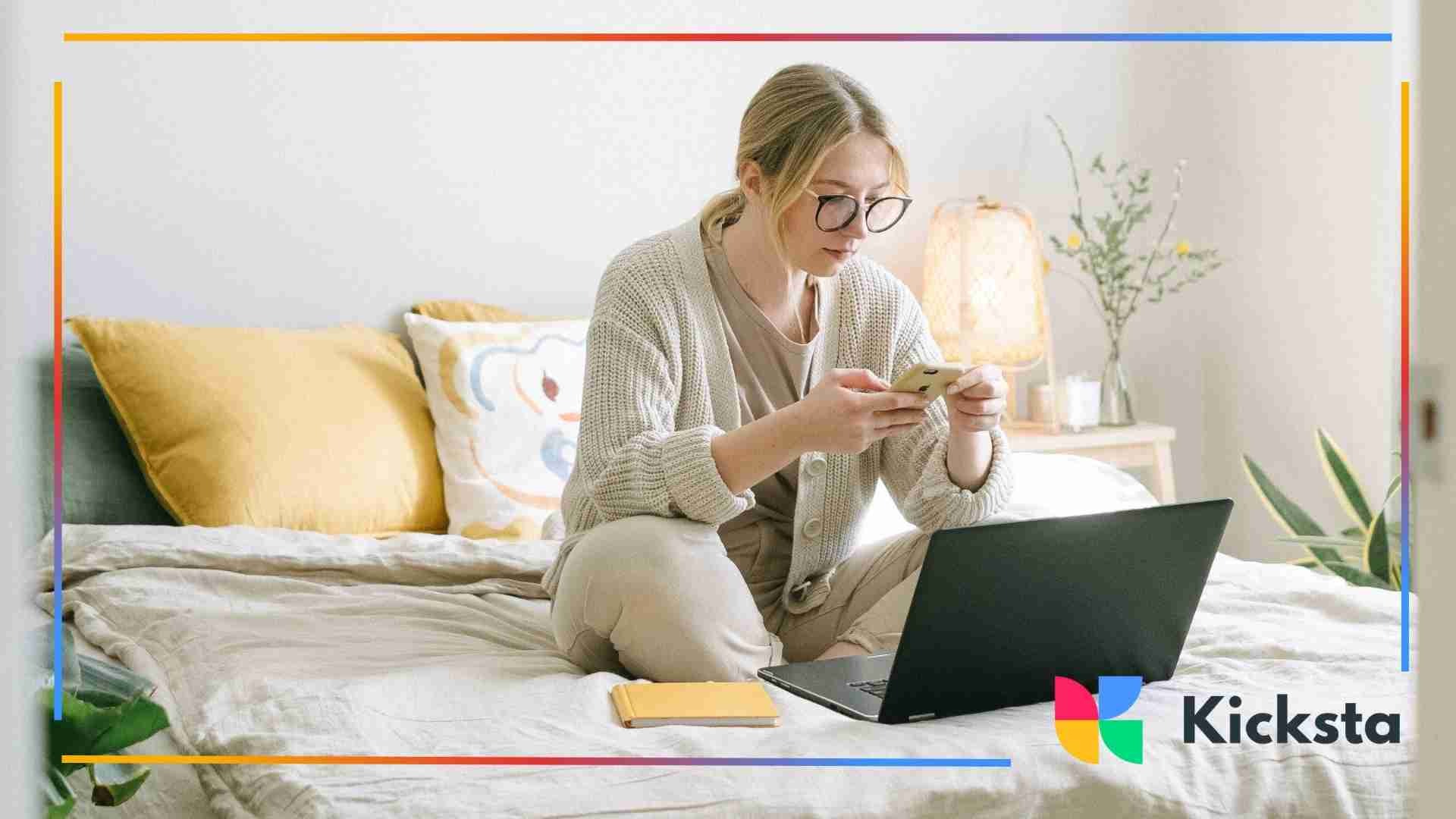 Woman sitting on her bed using her phone with an open laptop and notebook beside her.