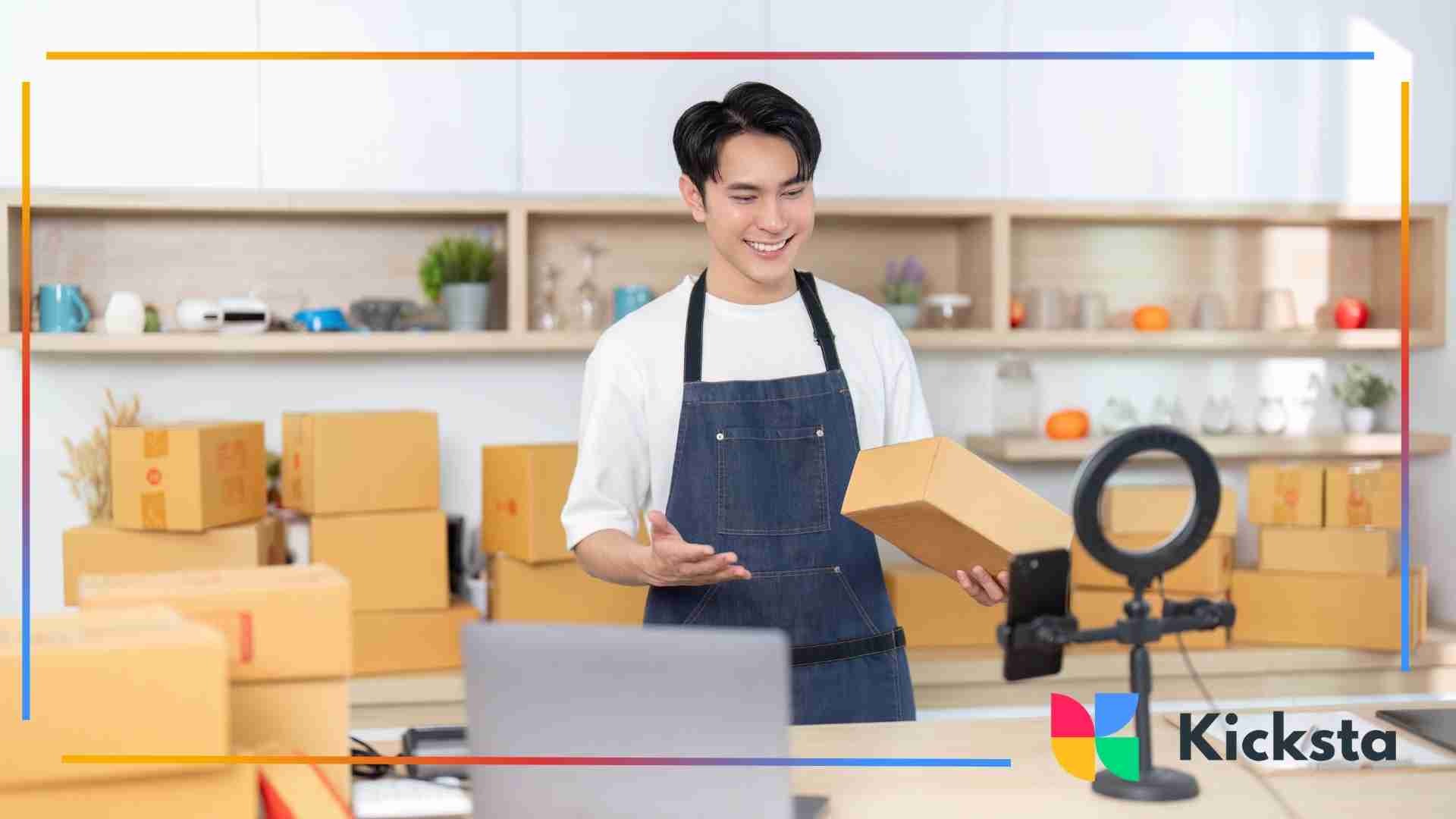 A small business owner wearing an apron records a product video using a smartphone and ring light while holding a shipping box in a workspace filled with packaging.