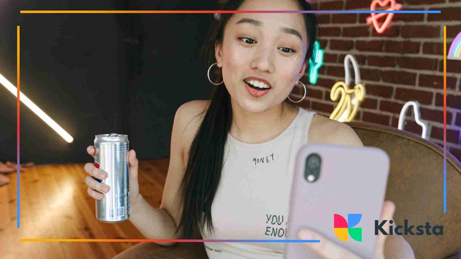 A young woman holding a can and smiling films herself with a smartphone in a room decorated with neon lights.