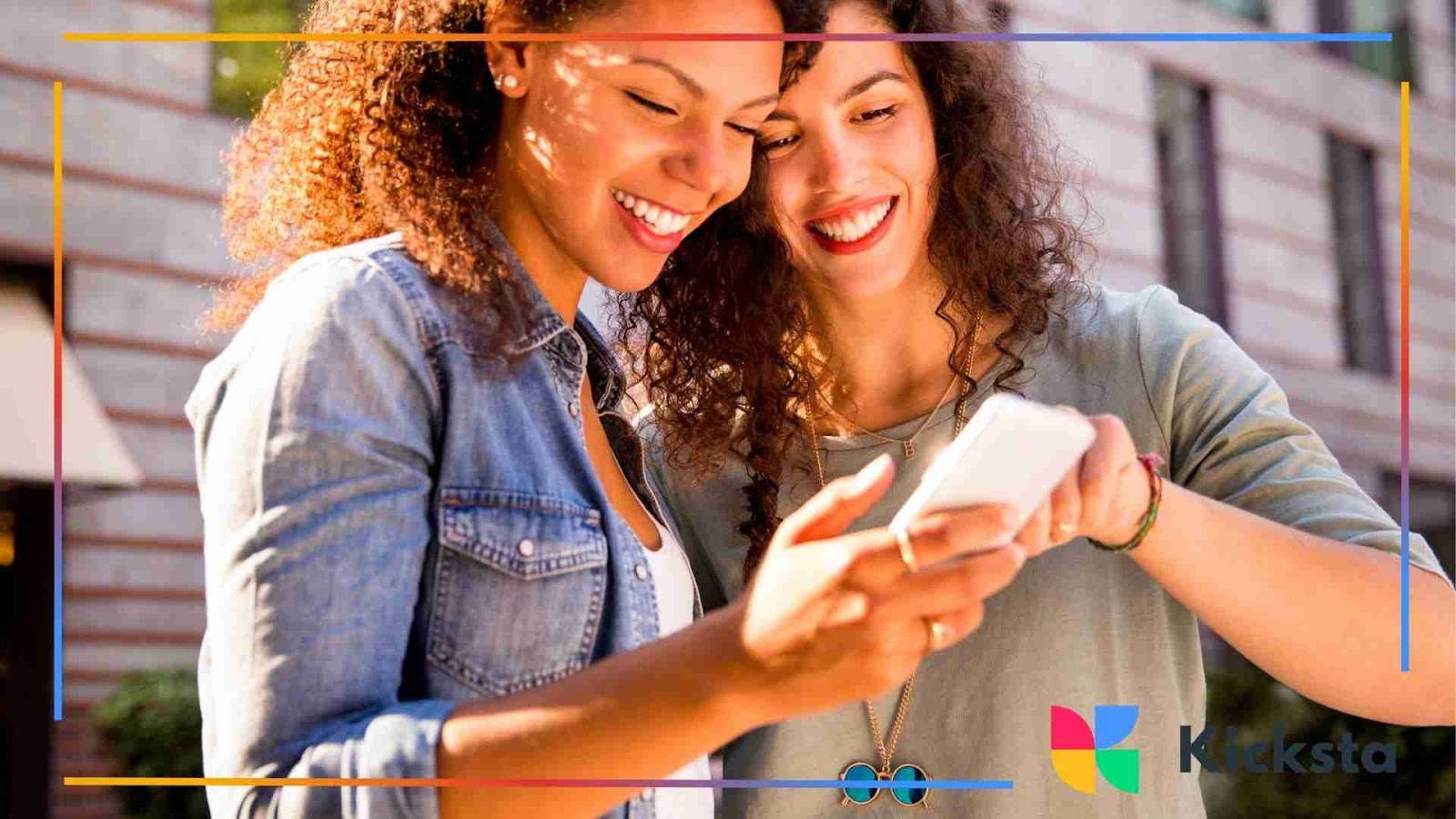 Two friends standing outdoors and smiling while looking at a smartphone together.
