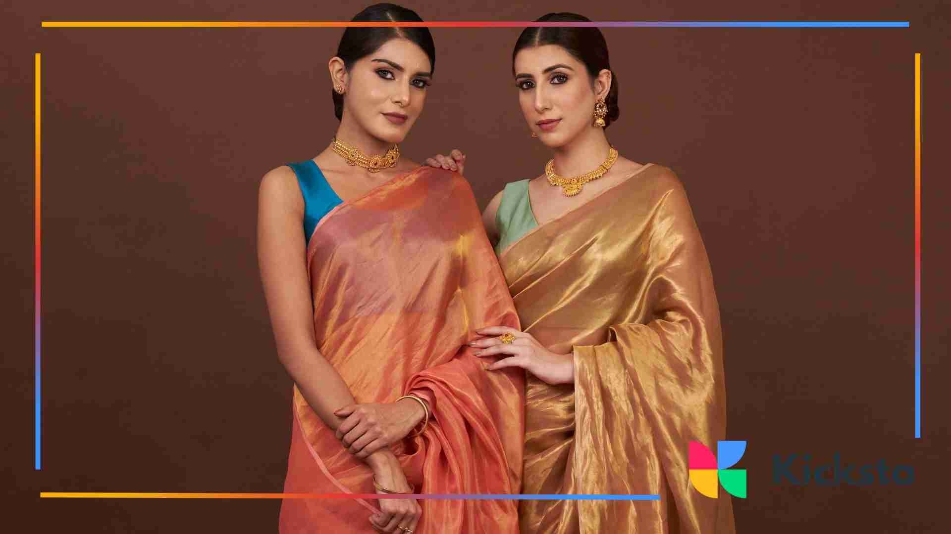Two women dressed in elegant silk sarees, one in orange and the other in gold, pose side by side against a brown background.